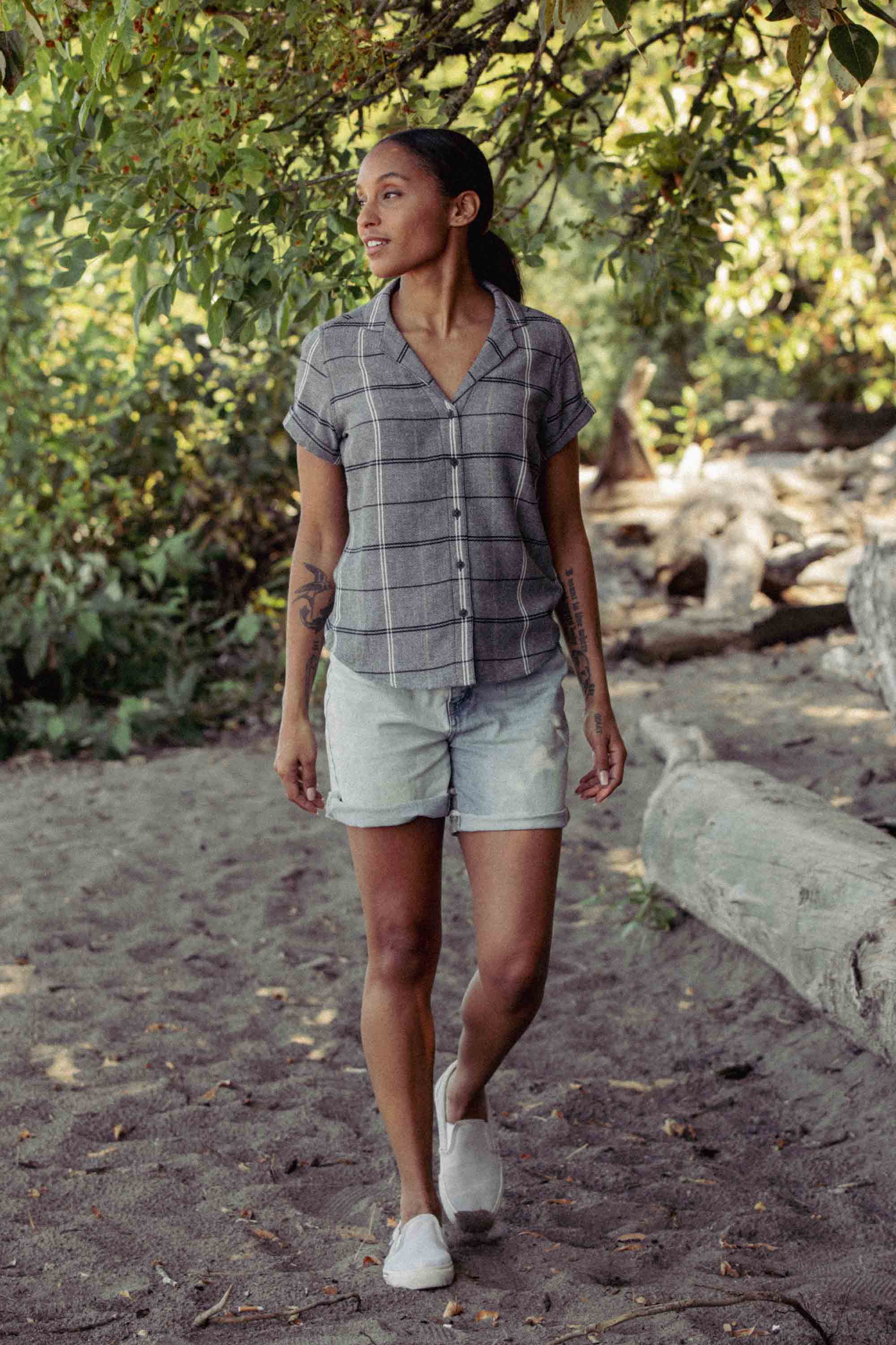 A person with tattoos on their arms walks outdoors in nature, wearing the Bridge & Burn Innes Shirt in Overcast Grid, paired with light denim shorts and white slip-on shoes as sunlight filters through the trees.
