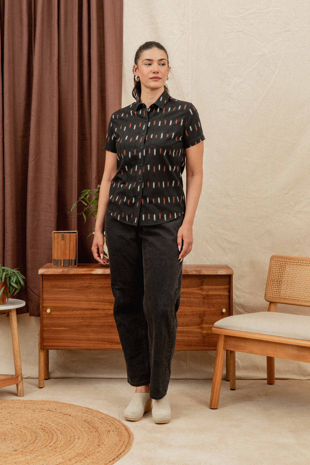 A woman stands indoors wearing the Bridge & Burn Lana Shirt in Dotted Horizon, a relaxed fit, short sleeve button-up made from 100% cotton, paired with black pants and white shoes. The shirt’s colorful pattern pops against the neutral room décor.