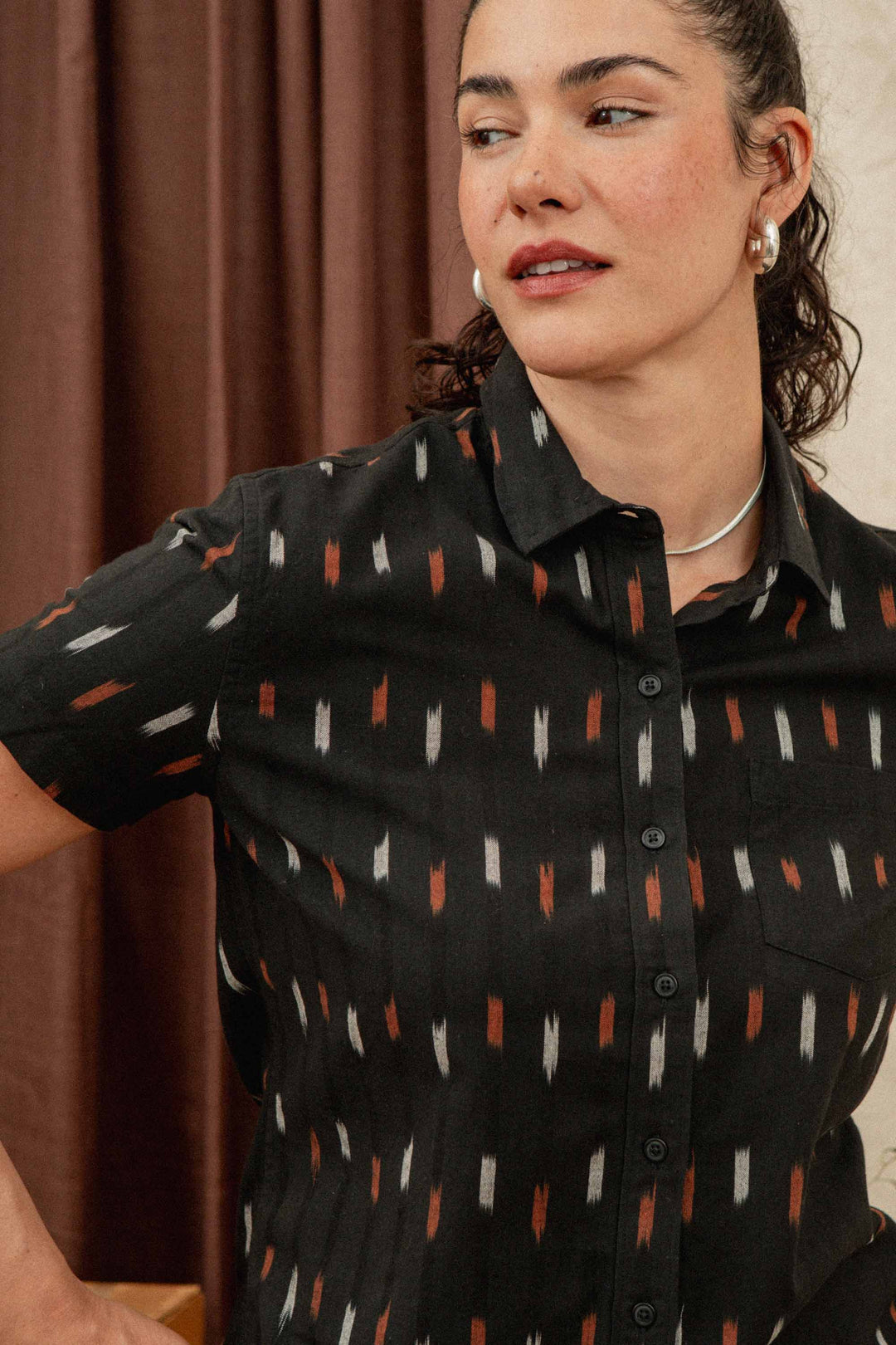 A woman with curly dark hair, wearing pearl earrings and the Bridge & Burn Lana Shirt in Dotted Horizon, stands before brown curtains and looks to her left.