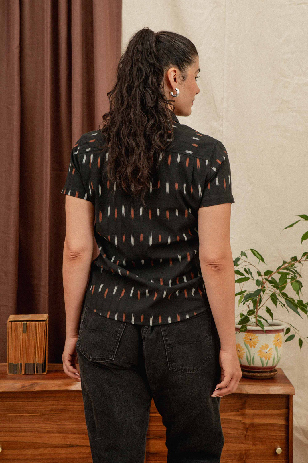 A woman with long, curly dark hair in a ponytail stands indoors with her back to the camera, wearing the Bridge & Burn Lana Shirt in Dotted Horizon and black jeans. A plant and wooden furniture are visible in the background.