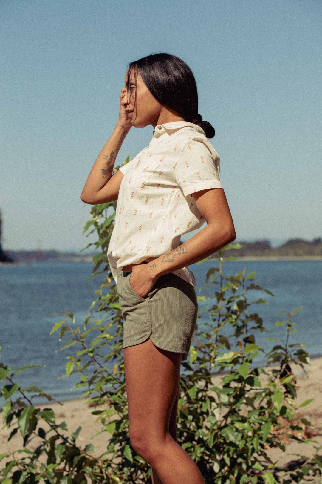 A woman stands outdoors by water and greenery on a sunny day, wearing the Bridge & Burn Lana Shirt in Dune Chevron with olive shorts, her hand touching her face and hair pulled back.