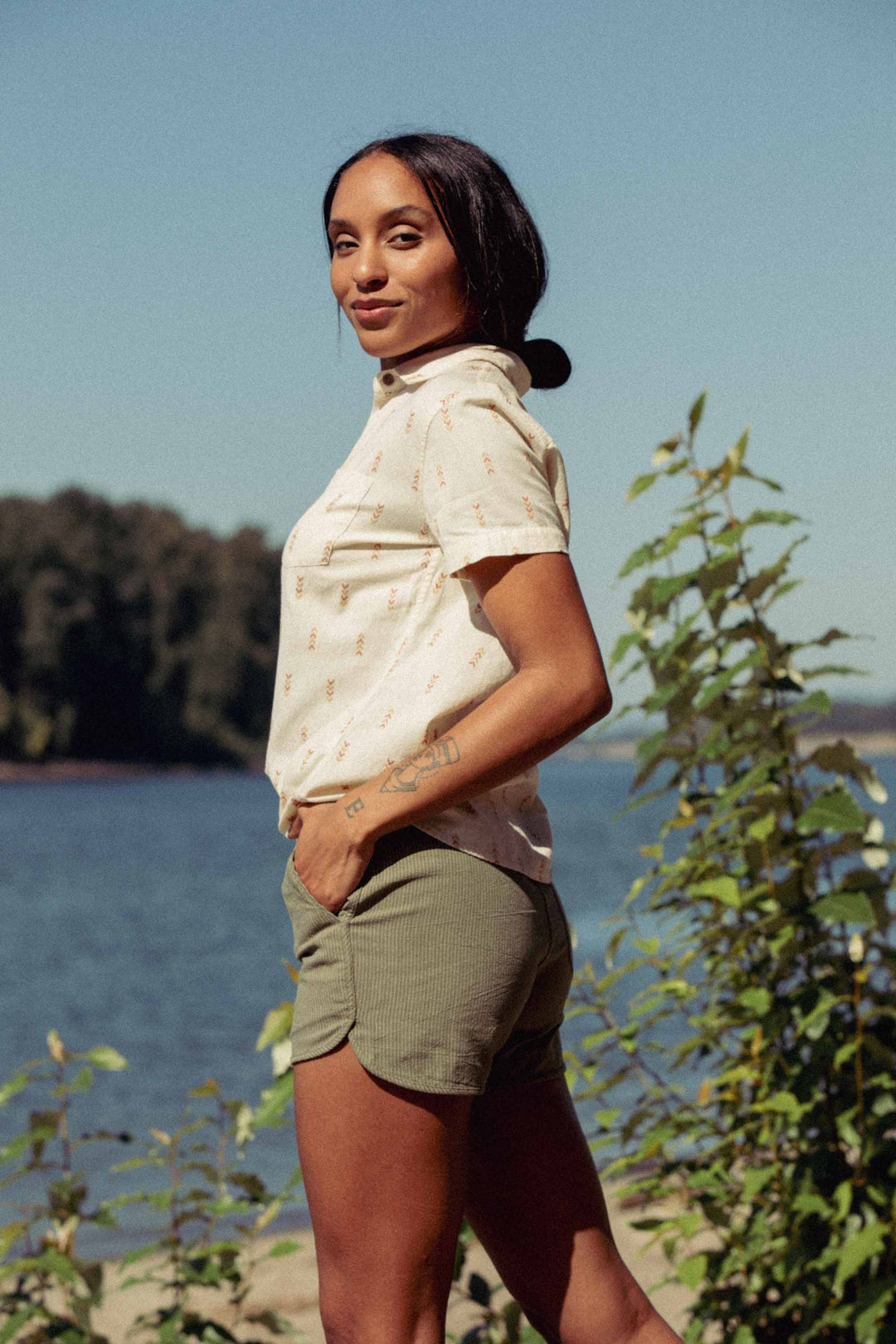 A woman stands outdoors by a river, wearing the Bridge & Burn Lana Shirt in Dune Chevron and green shorts, with her hair tied back. Green foliage and blue sky are visible in the background.