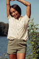 A woman stands outdoors by the water, smiling with arms raised. She wears the Bridge & Burn Lana Shirt in Dune Chevron and green shorts, with trees and blue sky behind her.