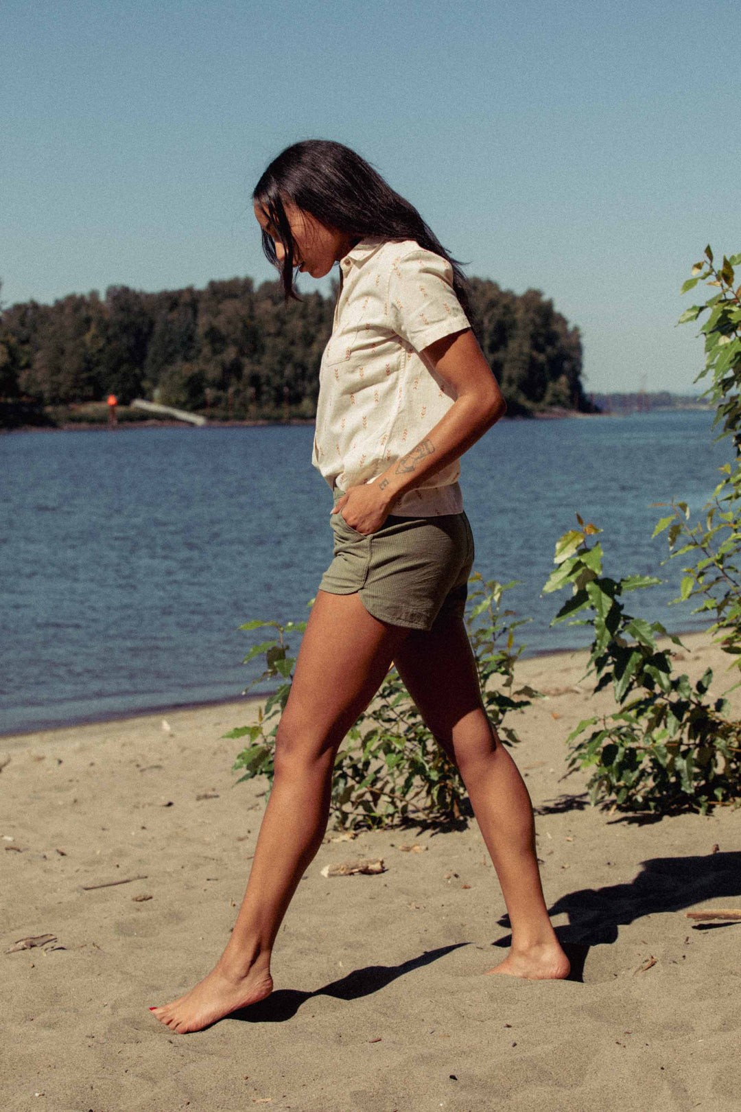 A person with long dark hair walks barefoot on the beach near water, wearing the Bridge & Burn Lana Shirt in Dune Chevron and olive green shorts. Green plants and trees appear in the background beneath a clear sky.