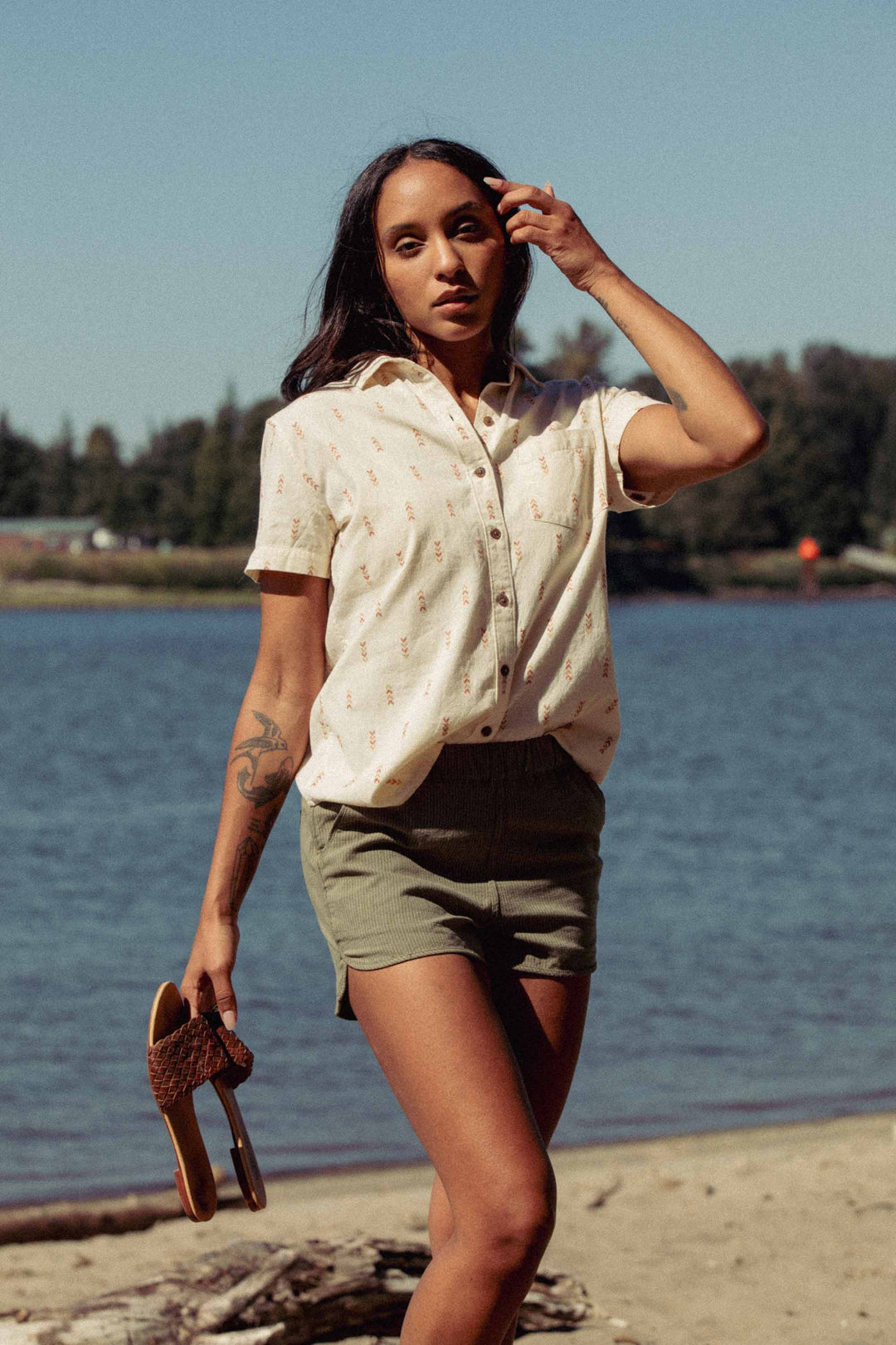 A person with long dark hair stands on a sandy beach near the water, wearing the Bridge & Burn Lana Shirt in Dune Chevron and green shorts, holding sandals in one hand and brushing their hair back. Trees and blue sky fill the background.