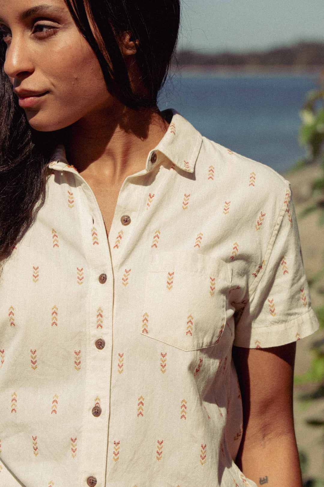 A woman with long dark hair wears the Bridge & Burn Lana Shirt in Dune Chevron, a 100% cotton short sleeve button-up, standing outdoors by water with blurred trees and sky behind her.