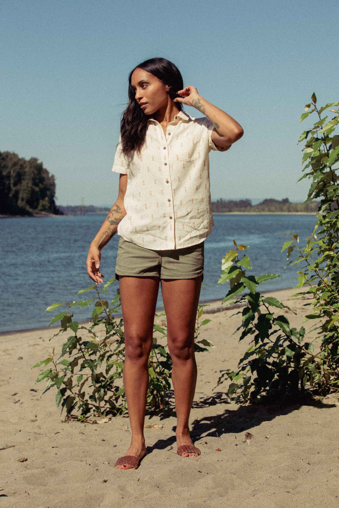 A woman with long dark hair and tattoos stands barefoot on a sandy beach near water, surrounded by greenery, wearing the Bridge & Burn Lana Shirt in Dune Chevron and olive shorts under a clear sky.