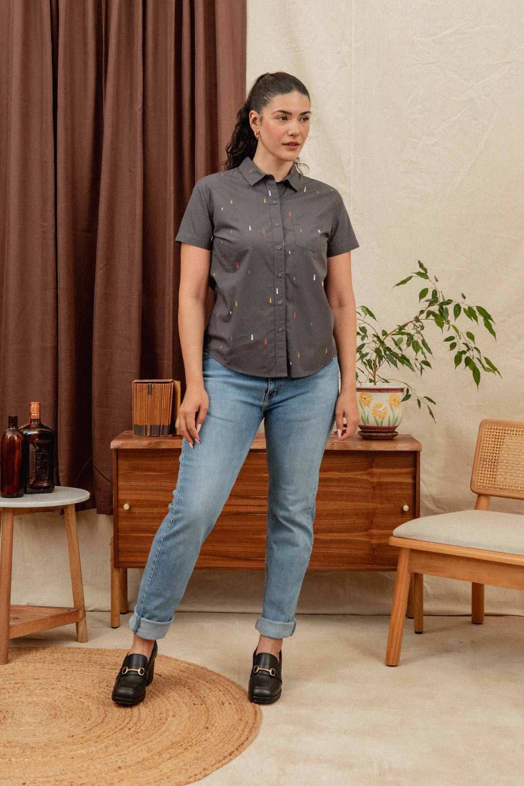 A woman with dark hair in a ponytail wears the Bridge & Burn Lana Shirt in Midnight Rain and blue jeans, standing on a beige rug in a cozy room with mid-century furniture, plants, and brown curtains.