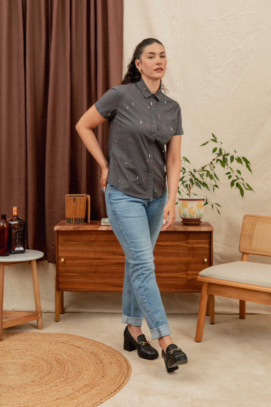 A woman with long dark hair wears the Bridge & Burn Lana Shirt in Midnight Rain, a 100% cotton relaxed fit short sleeve button-up, paired with light blue jeans. She stands indoors by a mid-century modern wooden cabinet with plants and chairs nearby.