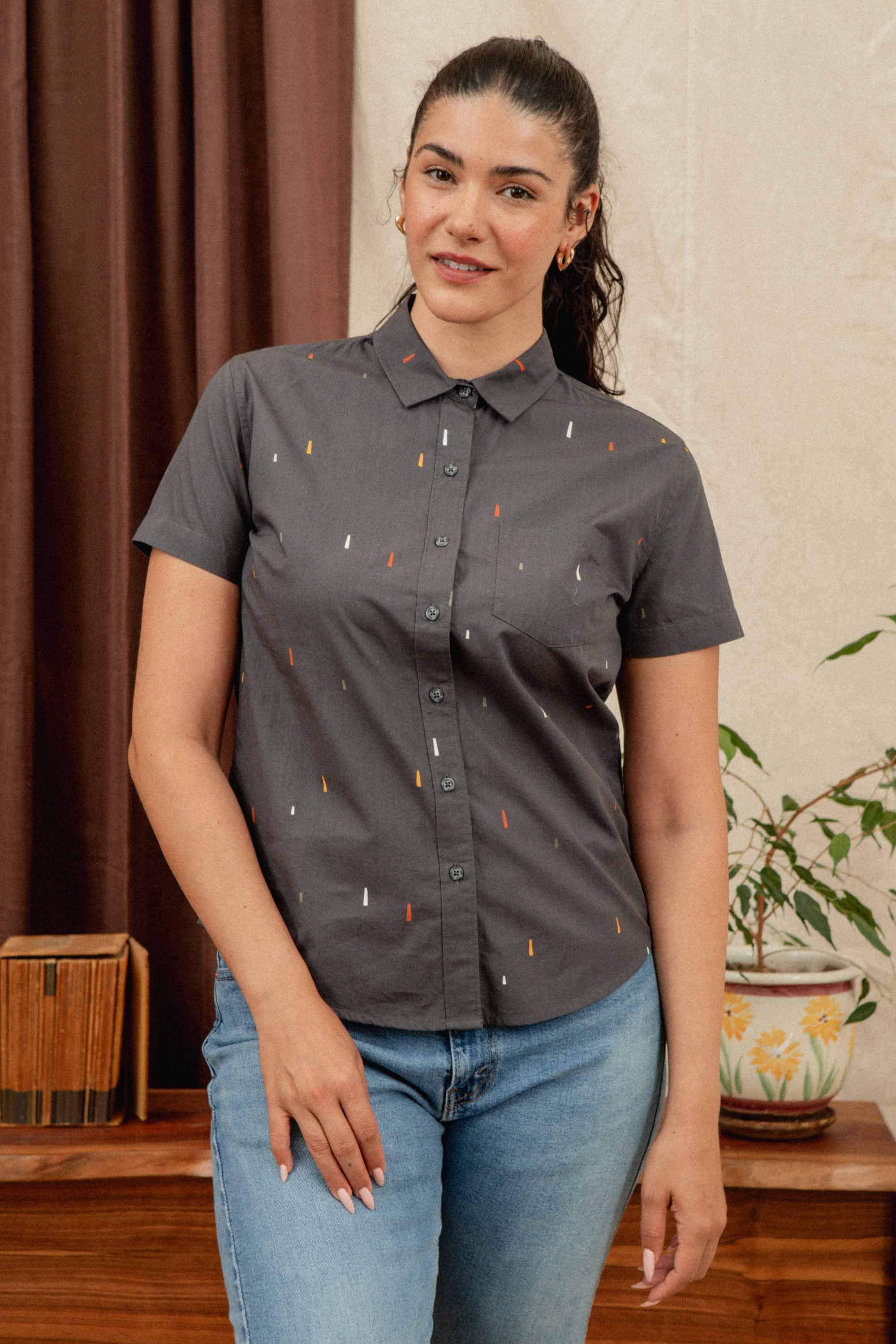 A woman with long dark hair in a ponytail wears the Bridge & Burn Lana Shirt in Midnight Rain—a relaxed fit, short sleeve button-up with a small colorful pattern—paired with light blue jeans, standing indoors beside a wooden table.