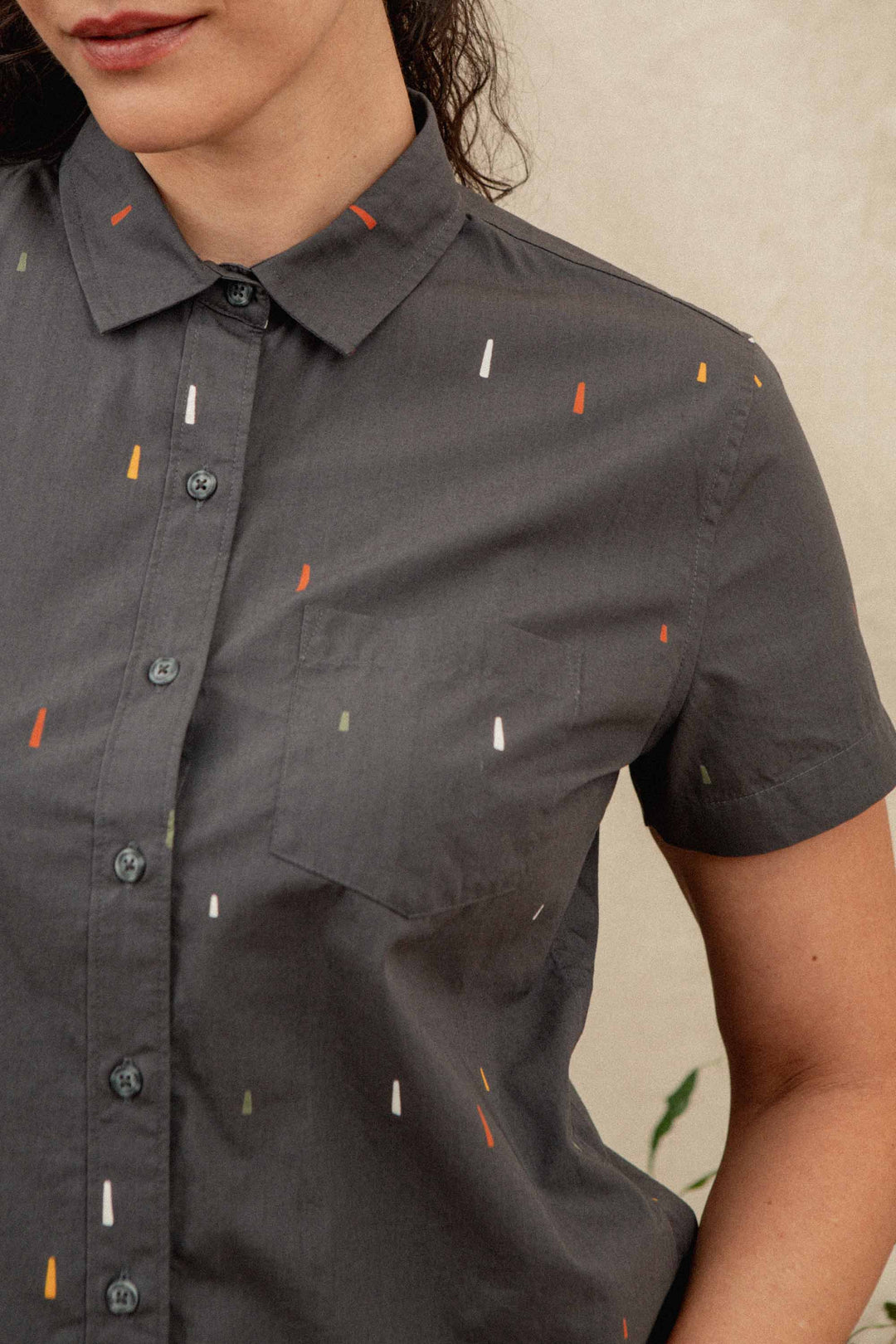 A woman is shown from the lower face to upper torso wearing the Bridge & Burn Lana Shirt in Midnight Rain, a relaxed fit short sleeve button-up in dark gray with small, colorful dash patterns.