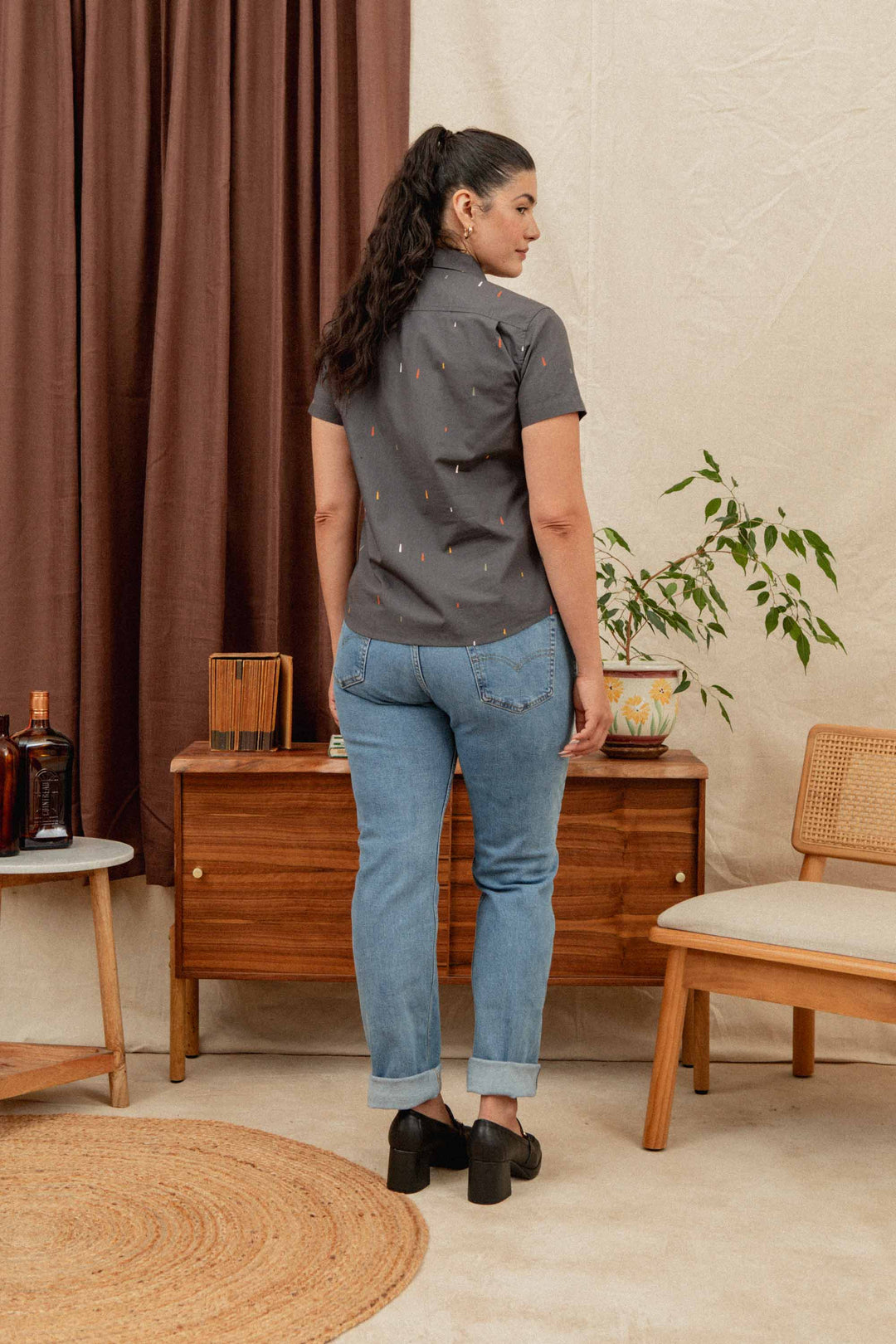 A woman with long dark hair in a ponytail stands indoors, wearing the Bridge & Burn Lana Shirt in Midnight Rain—a relaxed fit, short-sleeve gray 100% cotton top—paired with blue cuffed jeans and black shoes, amid mid-century furniture and potted plants.
