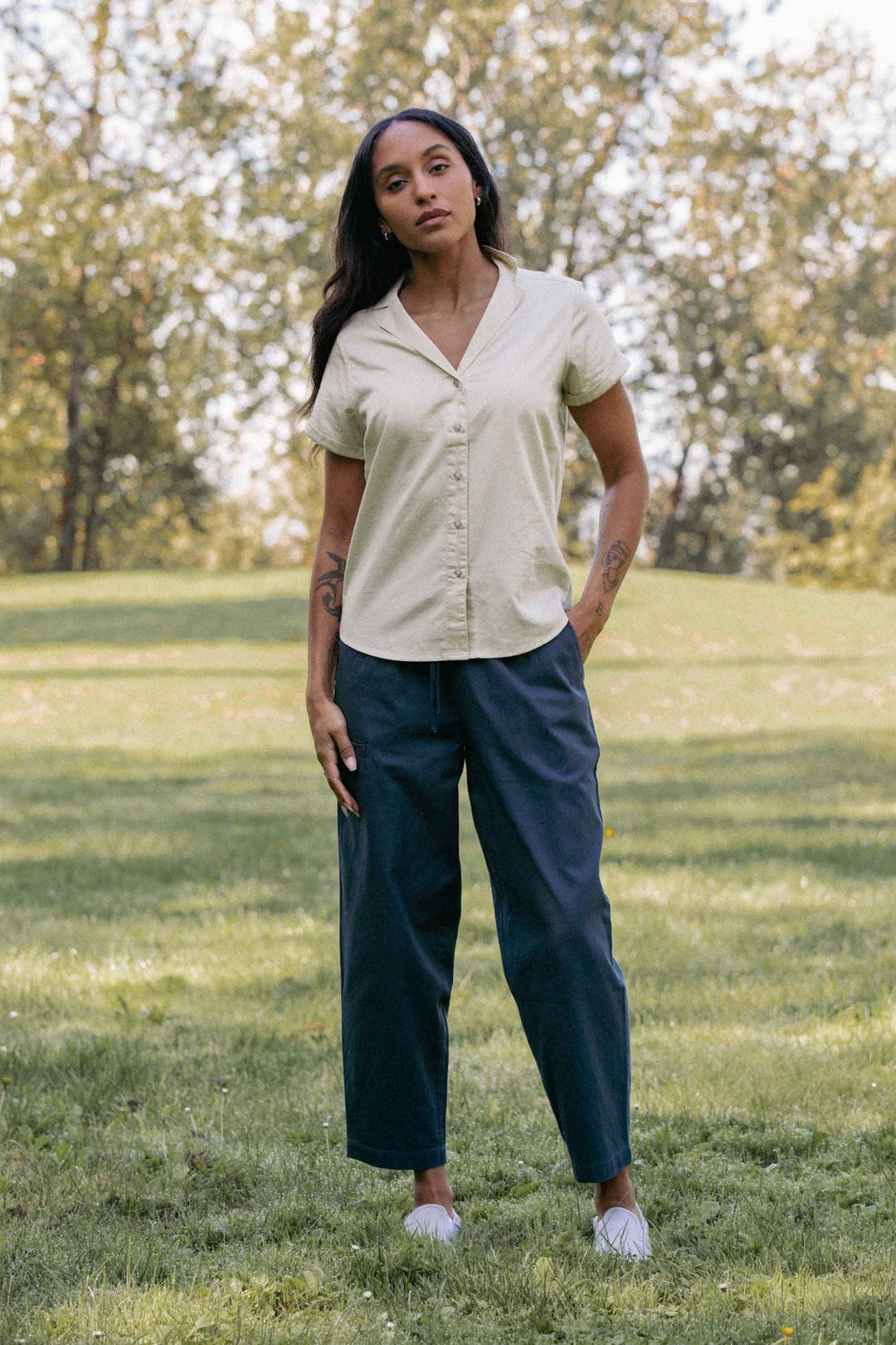 A woman stands on grass in a sunlit park, wearing the Bridge & Burn Lark Pant in Dark Slate with an elastic waist, paired with a short-sleeve beige button-up shirt. She has long hair and tattoos on her arms, surrounded by trees and a green lawn.
