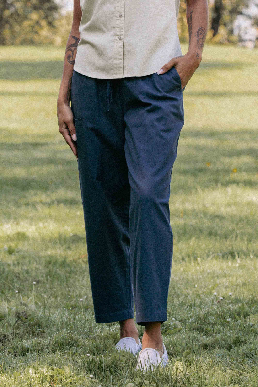 A person stands on grass with hands in pockets, wearing Bridge & Burn's Lark Pant in Dark Slate. Only their lower half is visible, showing the relaxed fit and elastic waist. Trees and greenery are softly blurred in the background.