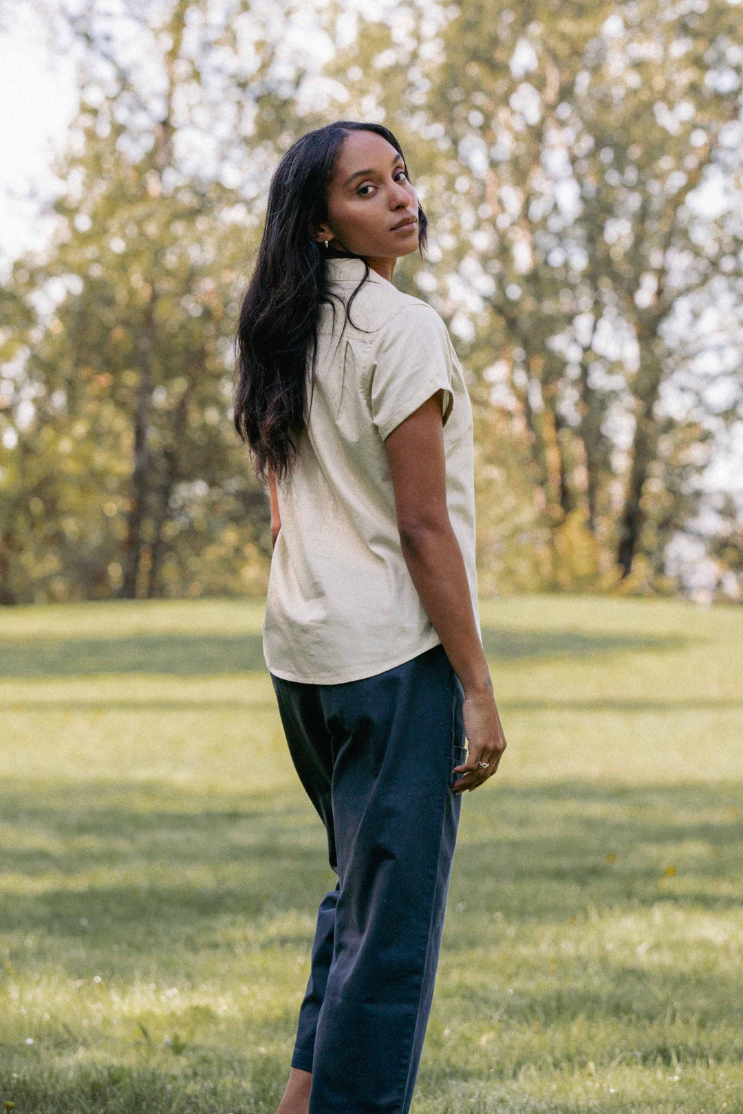 A woman with long dark hair stands on a grassy field, looking over her shoulder at the camera. She wears Bridge & Burn's Lark Pant in Dark Slate and a light short-sleeved shirt, with trees and sunlight in the background.