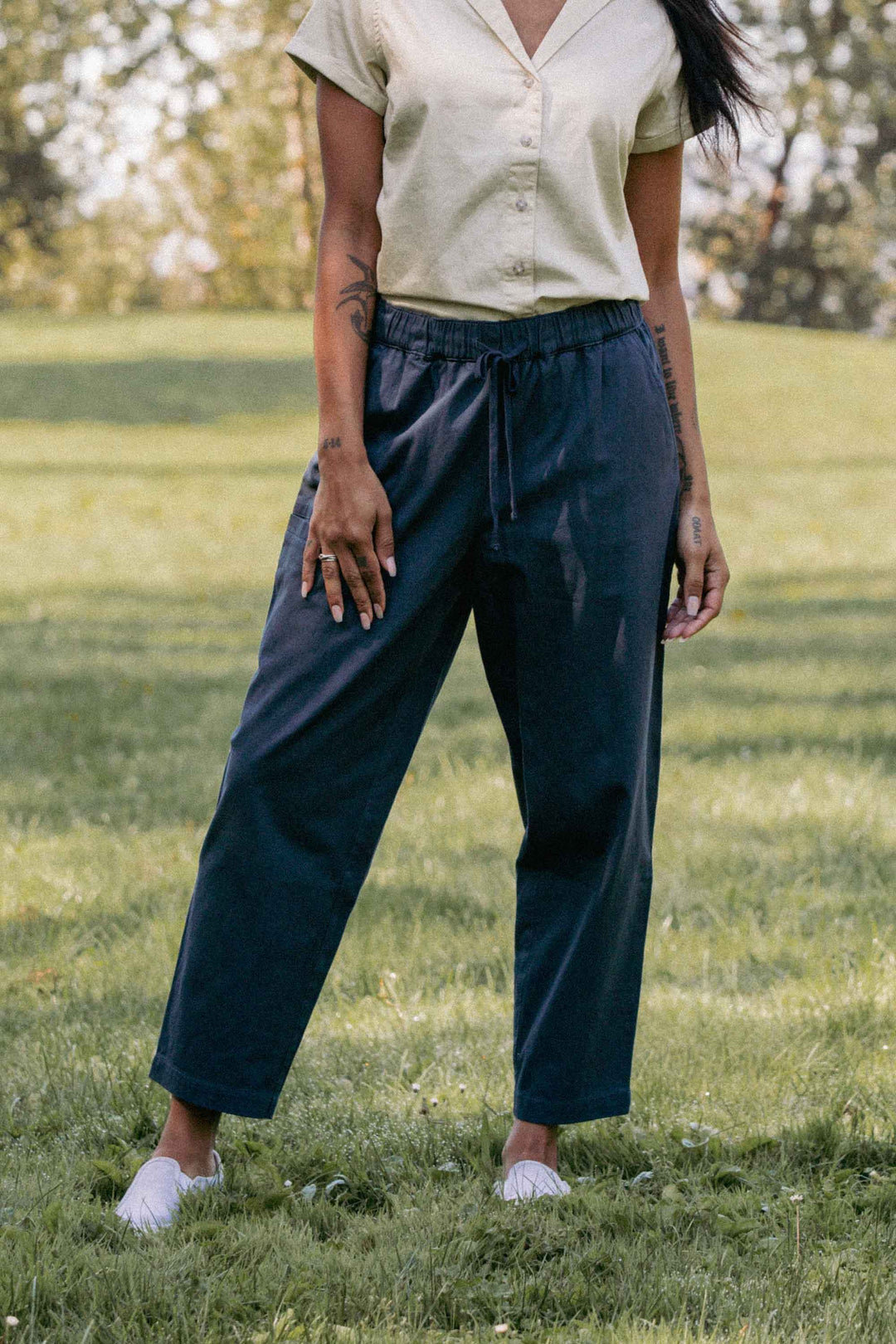 A person stands on grass in a sunny outdoor setting, wearing Bridge & Burn's Lark Pant in Dark Slate with an elastic waist, a short-sleeve light yellow shirt, and white shoes. Only their body from the shoulders down is visible.