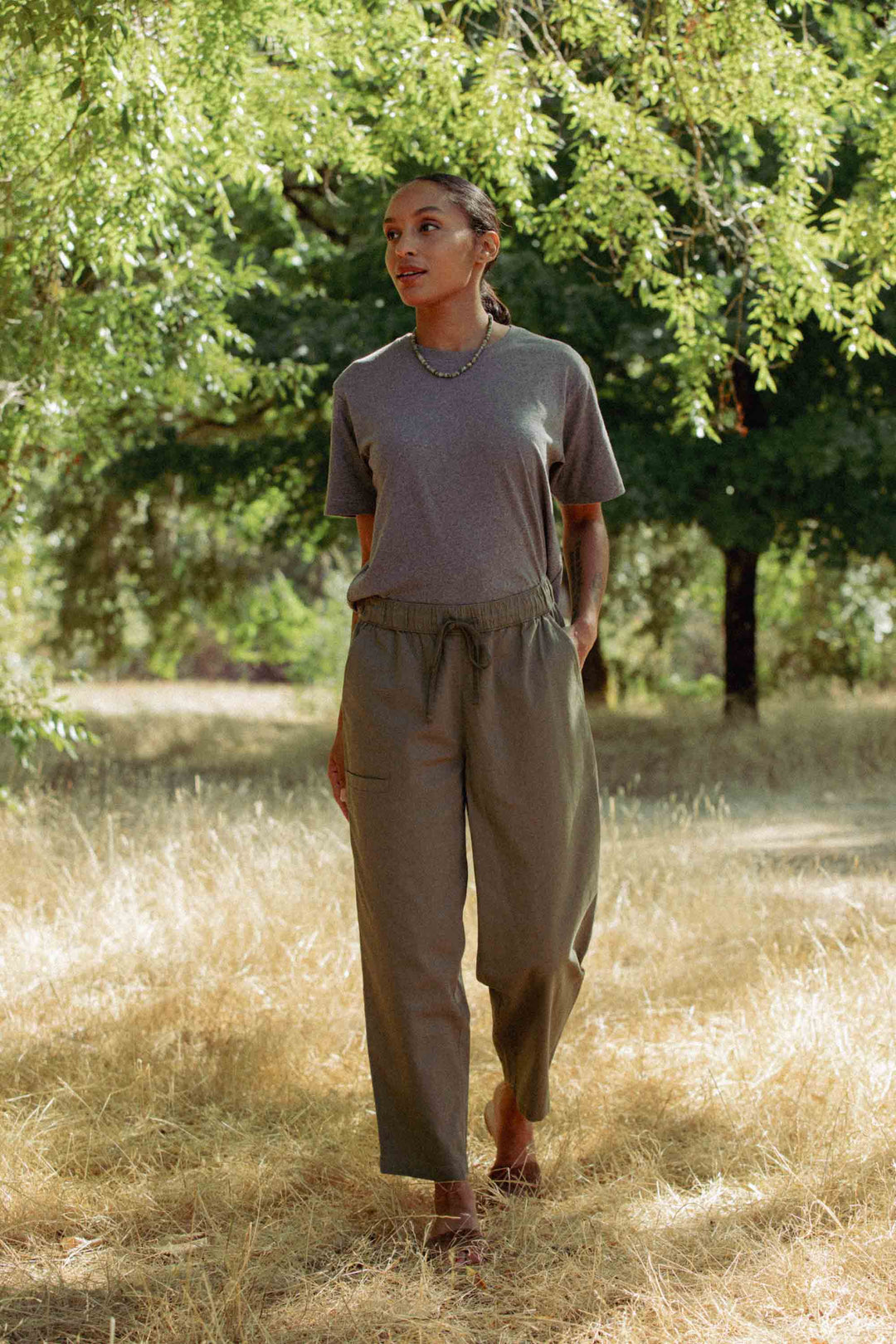 A person strolls through a sunlit grassy area beneath green trees, wearing a gray t-shirt and the Lark Pant in Olive by Bridge & Burn, hands in pockets, finished with a necklace.