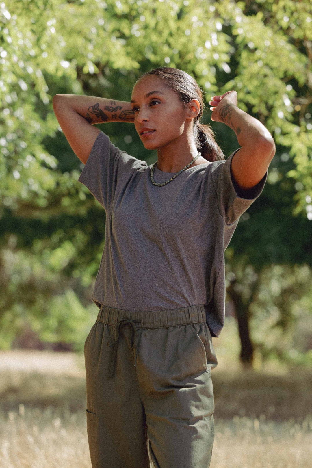 A tattooed person stands outdoors tying back their dark hair, wearing a gray t-shirt and Bridge & Burn's Lark Pant in Olive, along with a beaded necklace. Sunlight streams through the trees in the background.
