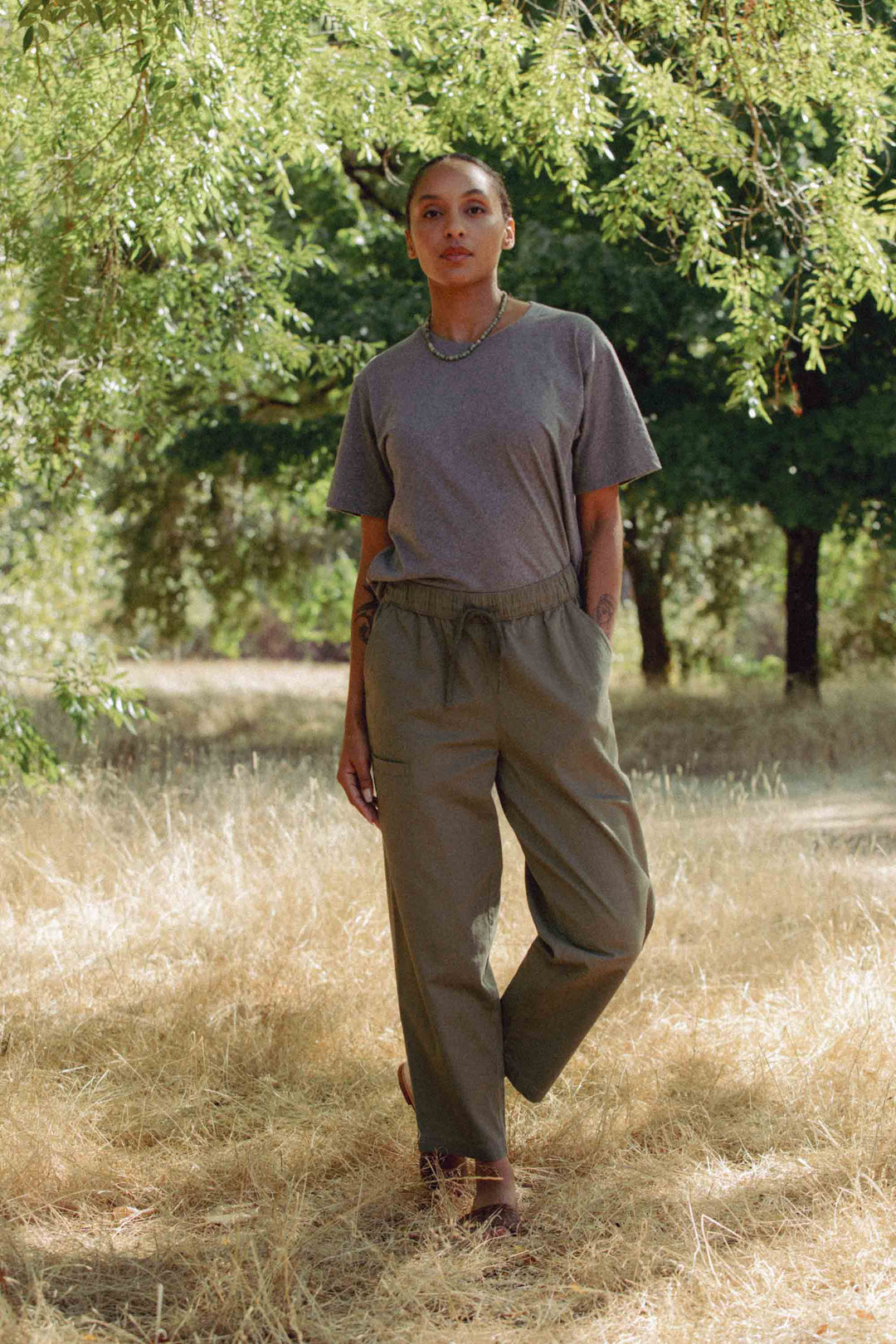 Standing outdoors on dry grass beneath leafy trees, a person wears Bridge & Burn’s Lark Pant in Olive and a gray t-shirt. Sunlight filters through the branches, creating a calm, natural atmosphere.