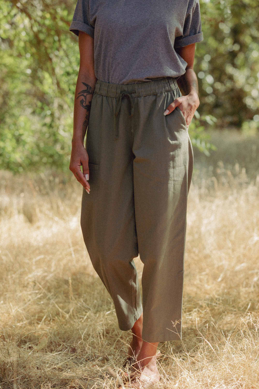 A person stands barefoot on dry grass, wearing Bridge & Burn's Lark Pant in olive with a relaxed fit and elastic waist, plus a gray short-sleeve shirt. A tattoo is visible on their right forearm against a backdrop of lush green foliage.