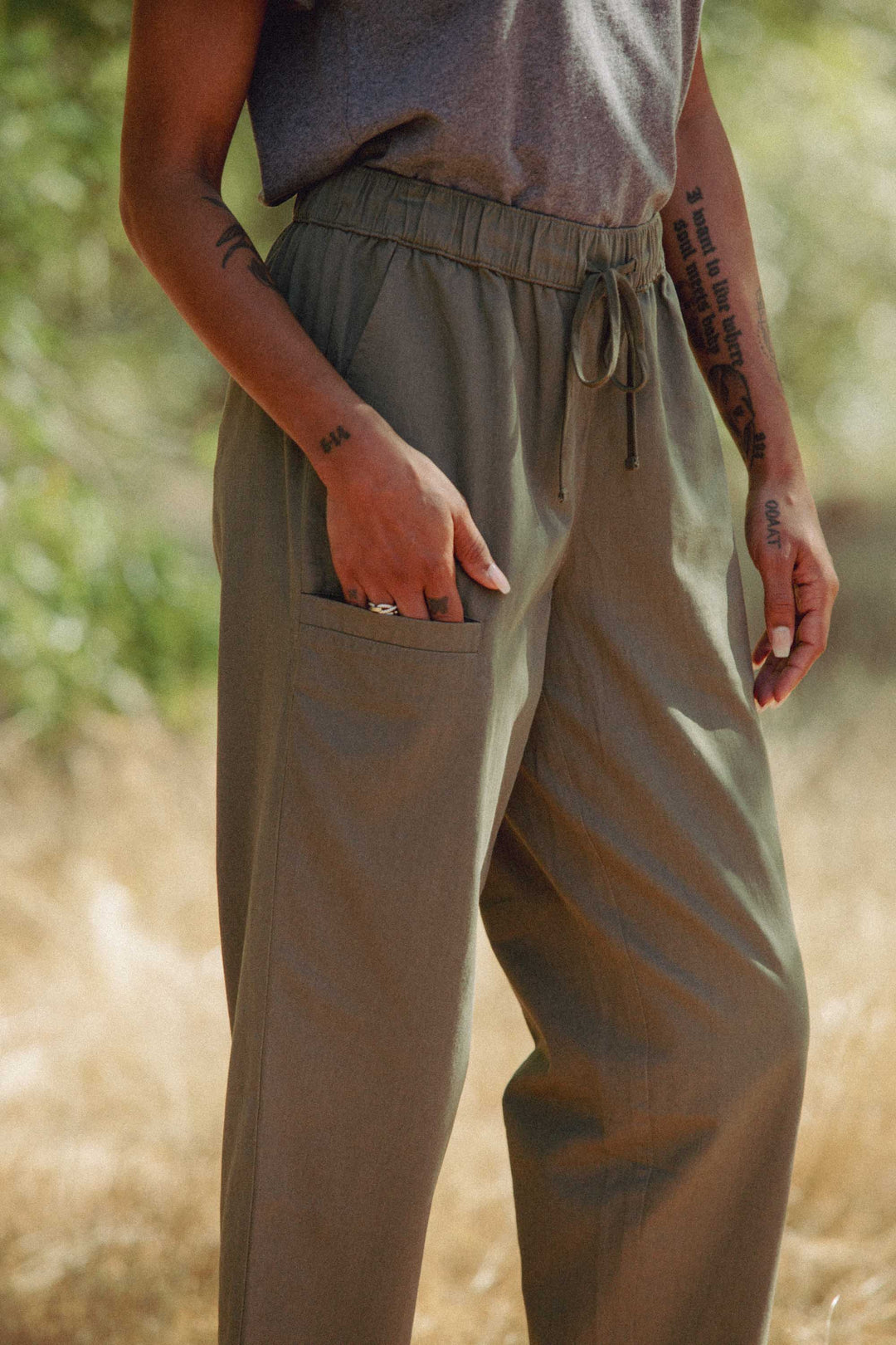 A person stands outdoors in Bridge & Burn’s Lark Pant in olive, featuring a relaxed fit and elastic waist, crafted from organic cotton, paired with a gray T-shirt. Tattoos are visible on their arms; the blurred background shows greenery and dried grass.