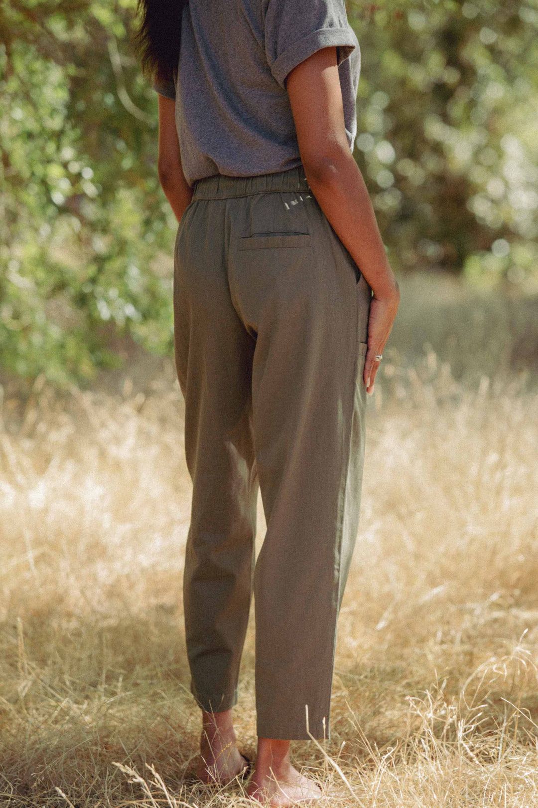 A person stands barefoot on dry grass, wearing Bridge & Burn's Lark Pant in olive and a gray t-shirt. The photo is taken from behind, outdoors with sunlit green foliage in the background.