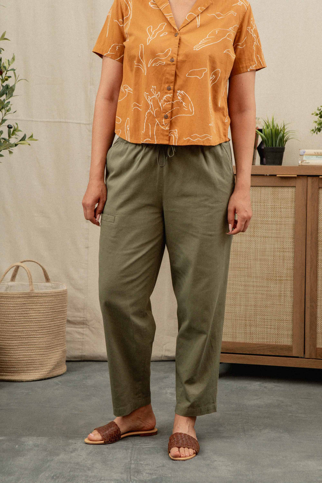 A person wearing an orange short-sleeve shirt, Bridge & Burn’s Lark Pant in Olive with an elastic waist, and brown woven slide sandals stands indoors near a rattan cabinet and plants.