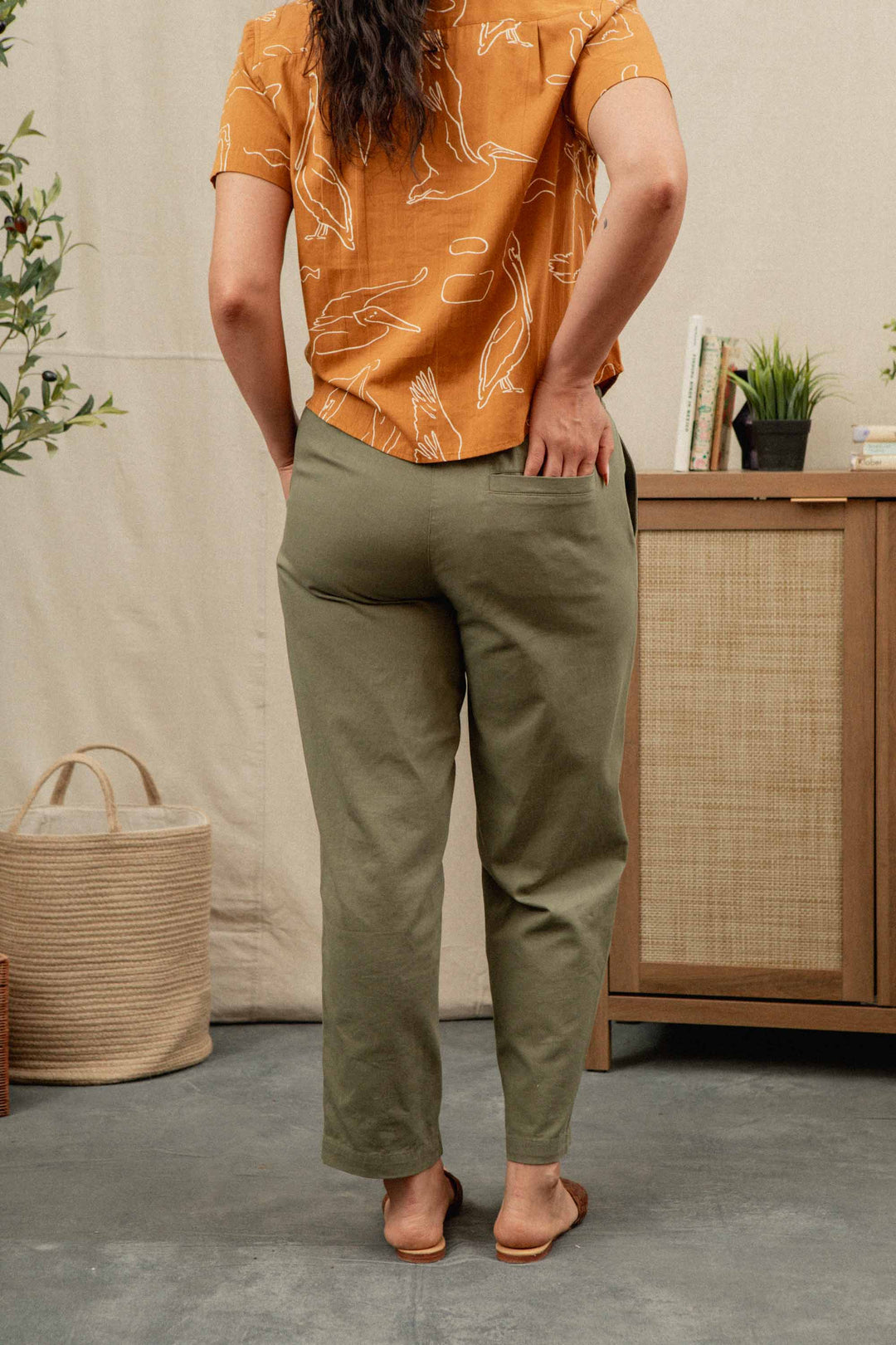 A person stands indoors, barefoot with one hand in their pocket, wearing the Bridge & Burn Lark Pant in Olive and an orange short-sleeve shirt. They're surrounded by plants and wooden furniture.