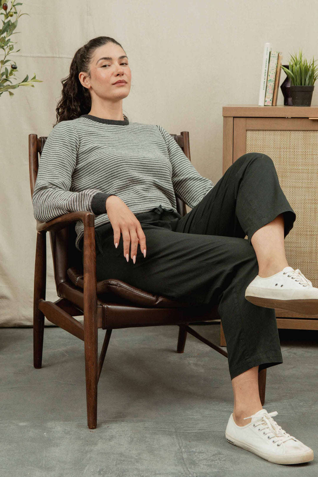 A woman with long dark hair sits confidently in a wooden chair, wearing a striped long-sleeve shirt, white sneakers, and Bridge & Burn’s Lark Pant in Soft Black. She rests one arm on the chair and crosses one leg over the other.