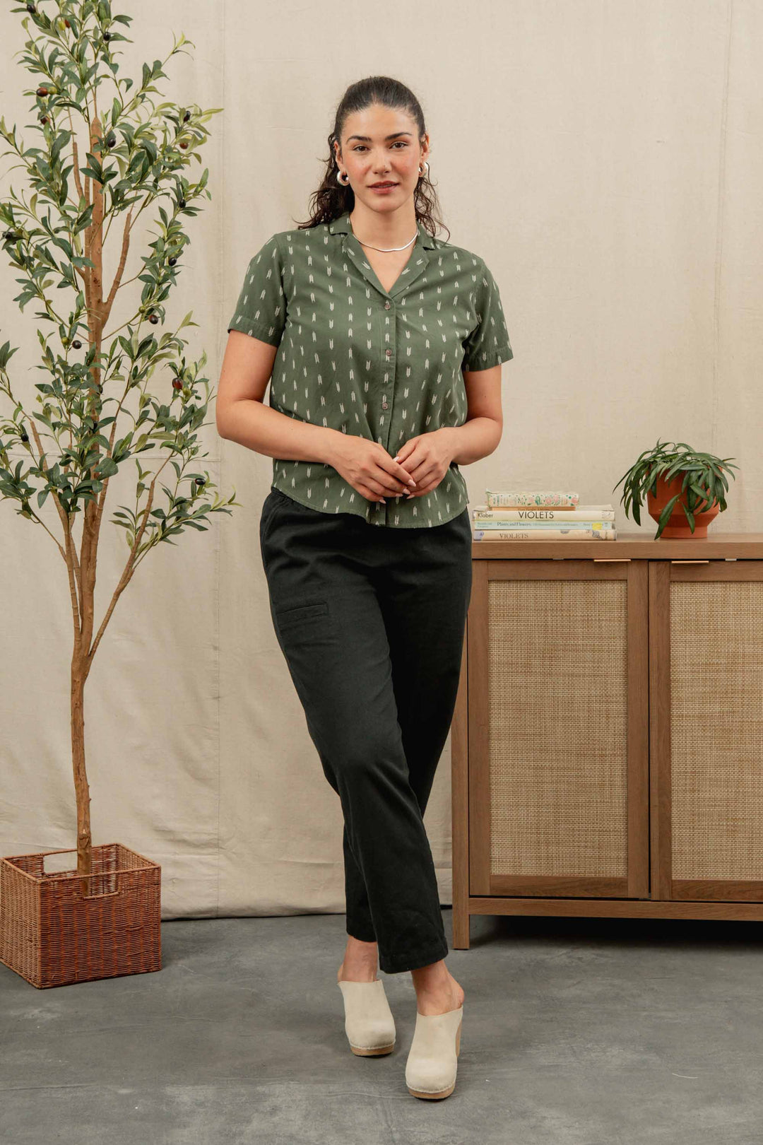 A woman stands indoors wearing relaxed fit Bridge & Burn Lark Pant in Soft Black, a green patterned short-sleeve shirt, and cream shoes. She is near a leafy potted plant and a wooden cabinet with books and a small plant.