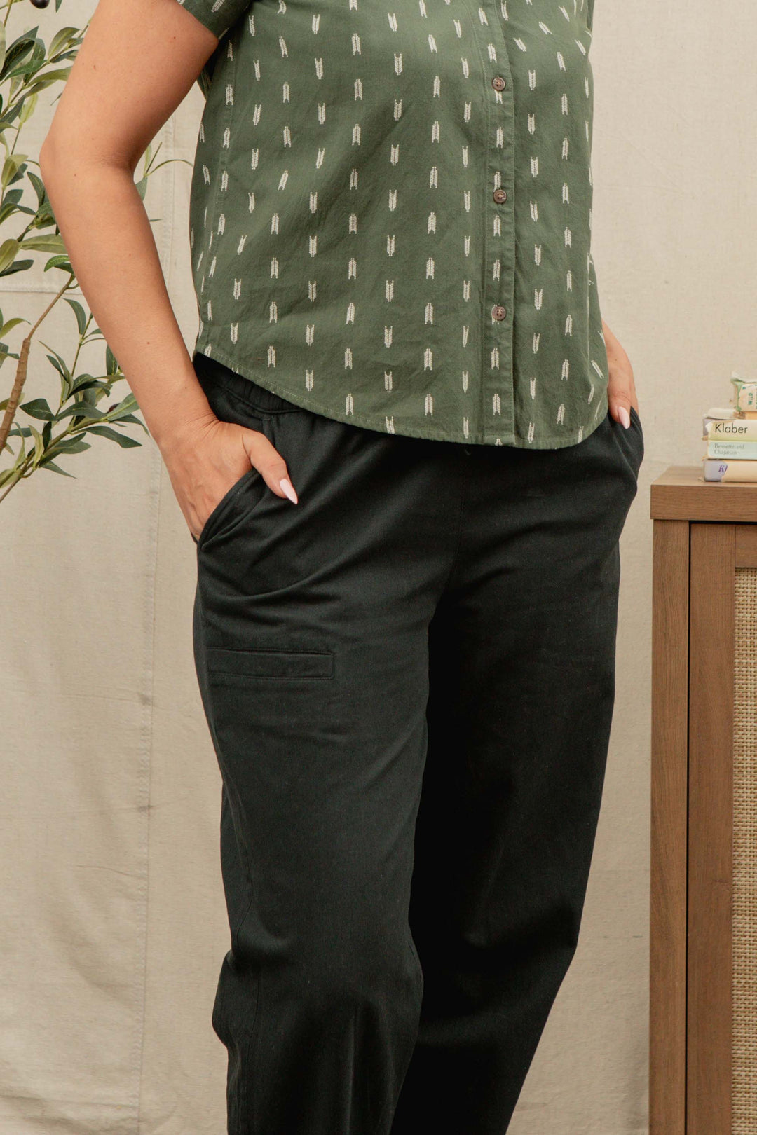 A person stands with hands in the pockets of Bridge & Burn's Lark Pant in Soft Black, wearing a short-sleeved green shirt with a small white pattern. Only the upper legs and torso are visible, with a plant and wooden cabinet behind.
