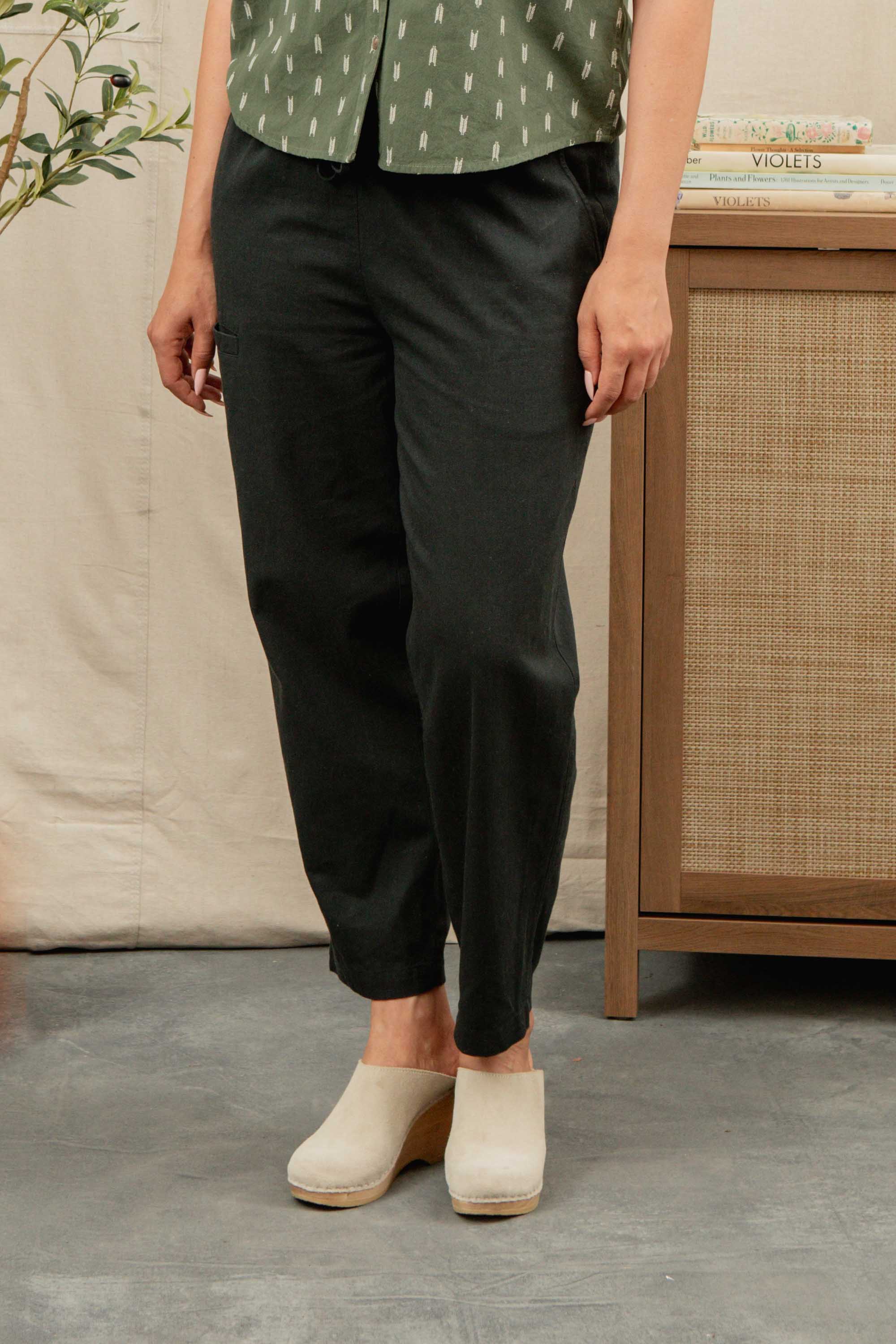 A person stands indoors wearing Bridge & Burn's Lark Pant in Soft Black with their hands at their sides; their head is not visible.