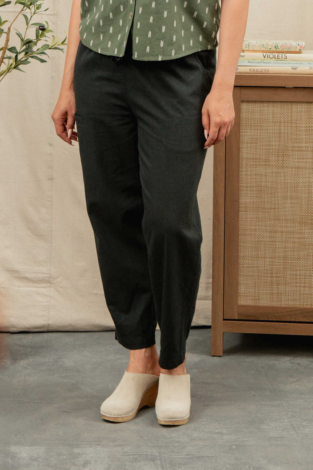 A person stands indoors wearing Bridge & Burn's Lark Pant in Soft Black with their hands at their sides; their head is not visible.