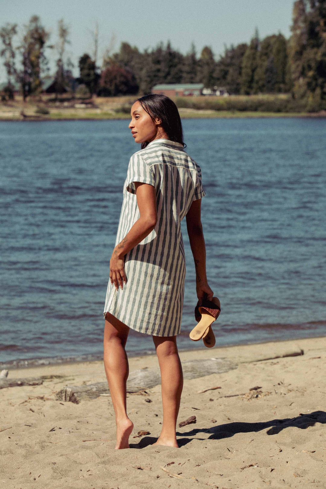 Barefoot on a sandy beach, a woman in the Bridge & Burn Loren Shirt Dress in Beach Stripe holds sandals and gazes over her shoulder toward the water and distant trees.
