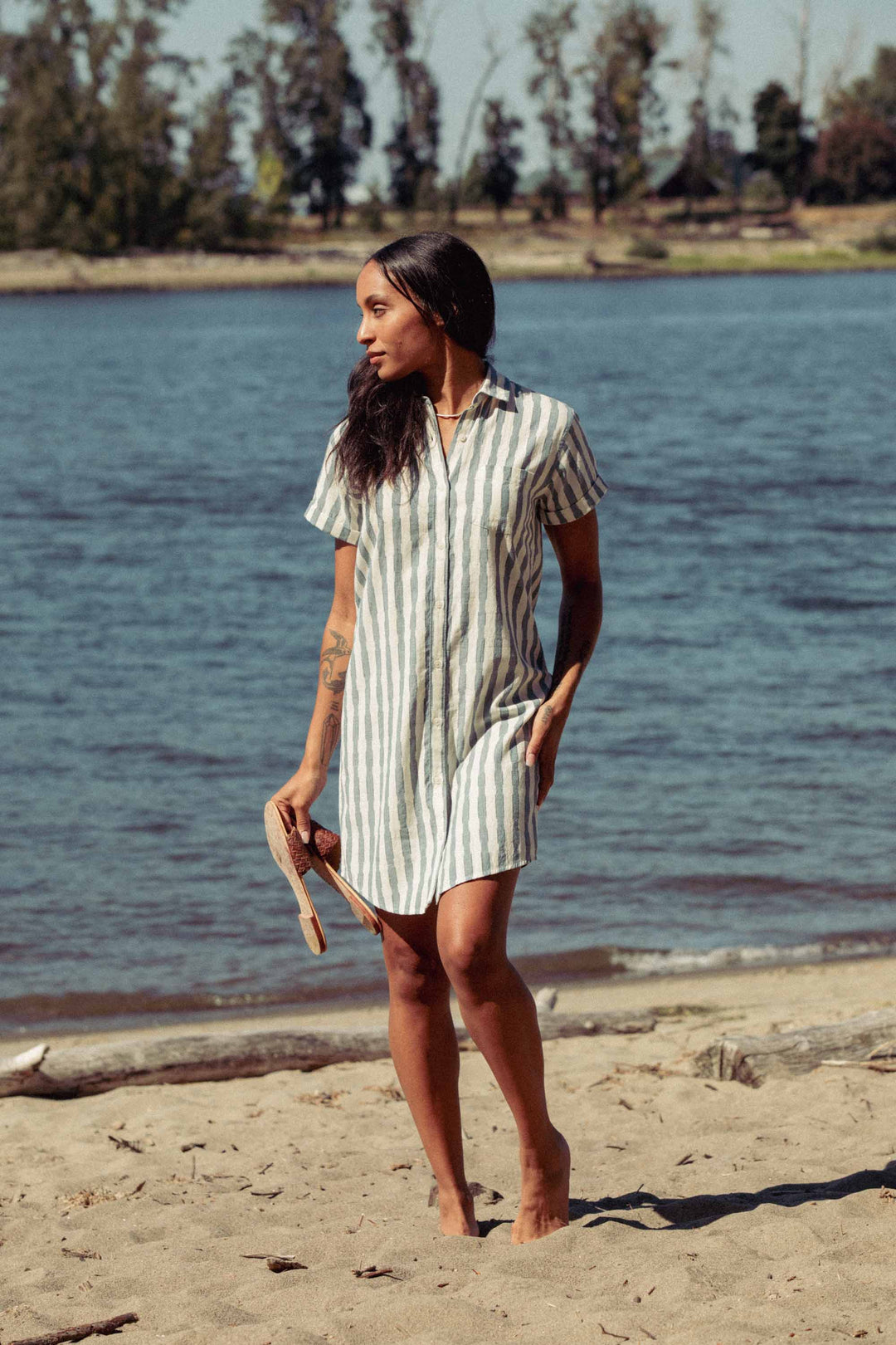 A woman in the Bridge & Burn Loren Shirt Dress in Beach Stripe stands barefoot on a sandy beach, holding sandals, with a river and trees in the background.