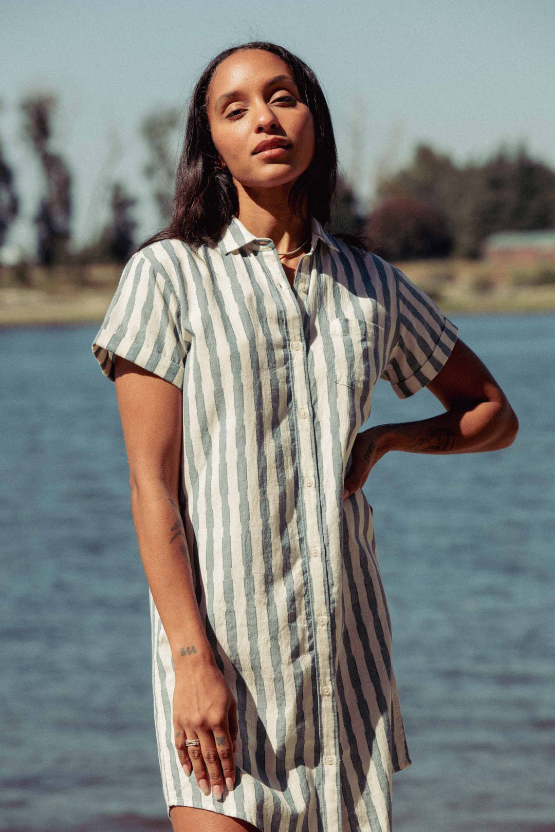 By a lakeside, someone confidently poses in the Bridge & Burn Loren Shirt Dress / Beach Stripe—a green and white 100% cotton button-front style with short sleeves, perfect for work or lounging amid a scenic backdrop.