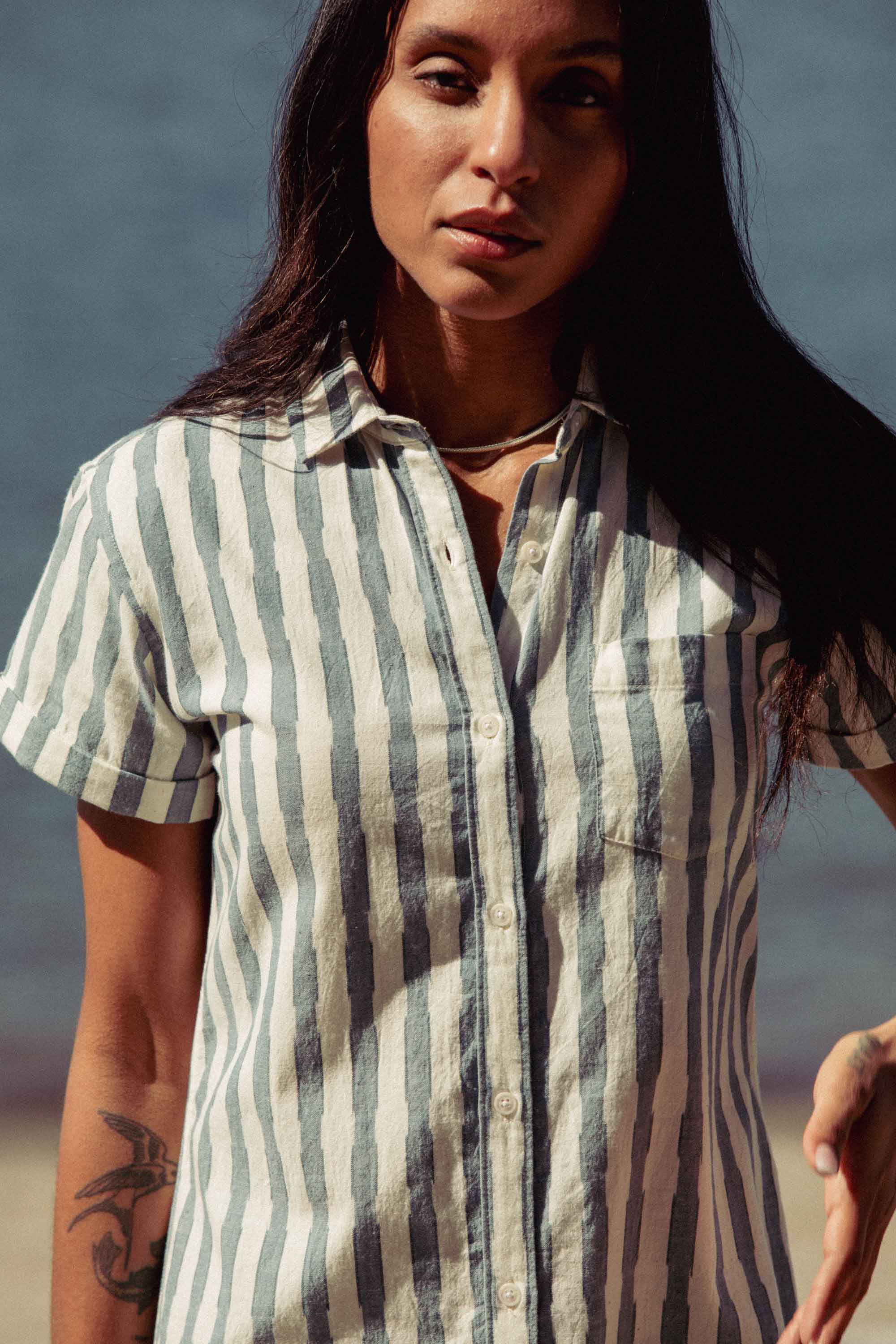 A woman with long dark hair and a tattoo on her left forearm stands outdoors by the water, wearing the Bridge & Burn Loren Shirt Dress in Beach Stripe— a short-sleeved, 100% cotton button-front dress with vertical stripes.