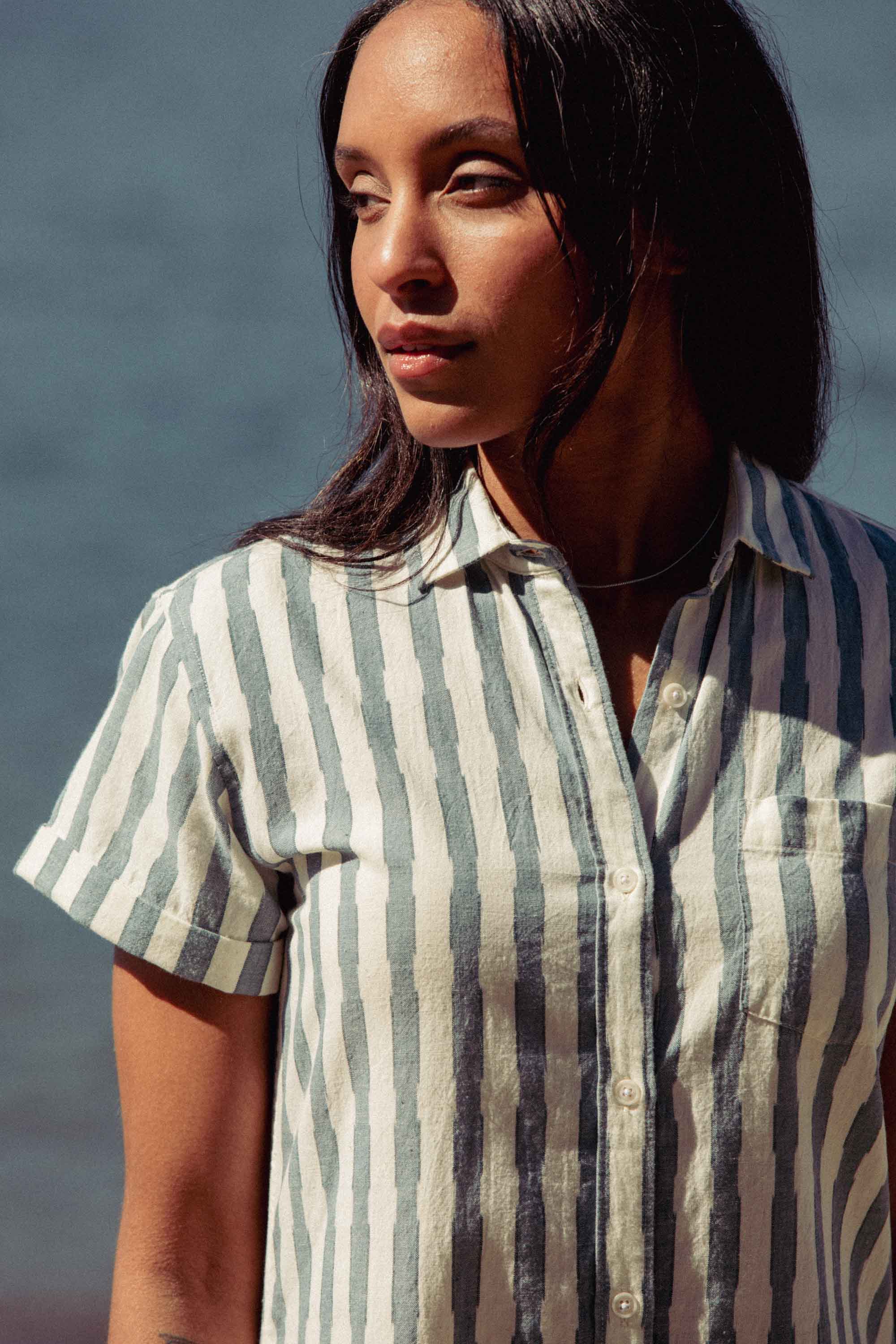 A woman with long dark hair wears the Bridge & Burn Loren Shirt Dress in Beach Stripe—a button front style with vertical white and gray stripes—as she stands outdoors by blurred water, gazing calmly to the side.