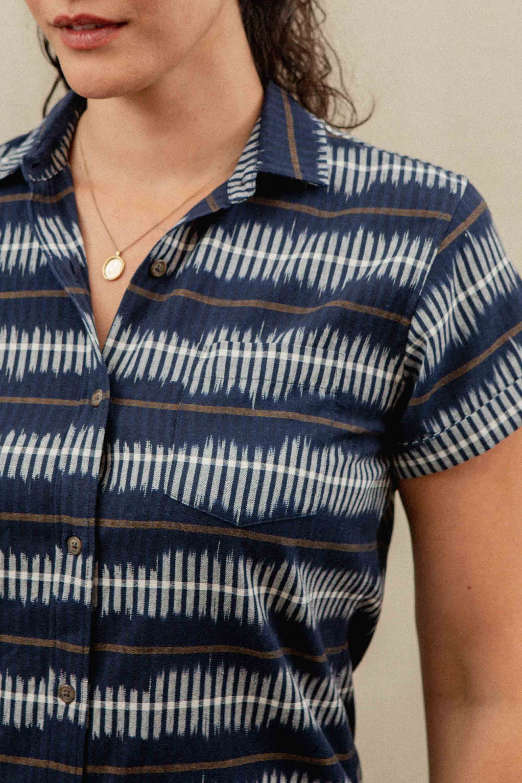 A woman wears the Loren Shirt Dress in Dusk Mirage by Bridge & Burn—a short-sleeved, blue button-up with white wavy stripes, made of 100% cotton. Only her lower face and upper torso are visible; she accessorizes with a pendant necklace.