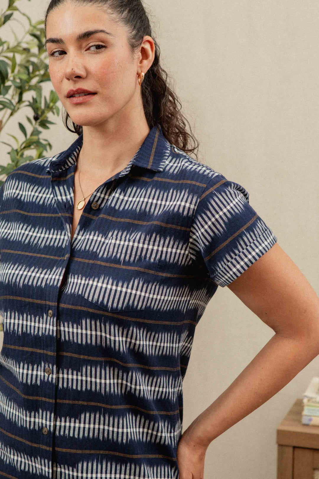 A woman with dark hair in a ponytail wears the Bridge & Burn Loren Shirt Dress in Dusk Mirage and gold jewelry, standing indoors by a potted plant and a wooden shelf with books, looking slightly to the side.