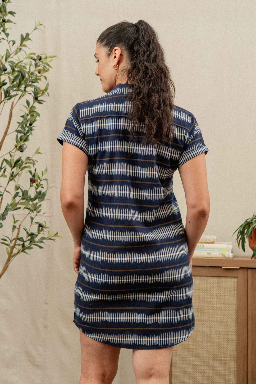 A woman with long curly hair in a ponytail stands indoors near a leafy plant and wooden cabinet, wearing the Bridge & Burn Loren Shirt Dress in Dusk Mirage—a short-sleeved, blue and white striped 100% cotton work dress.