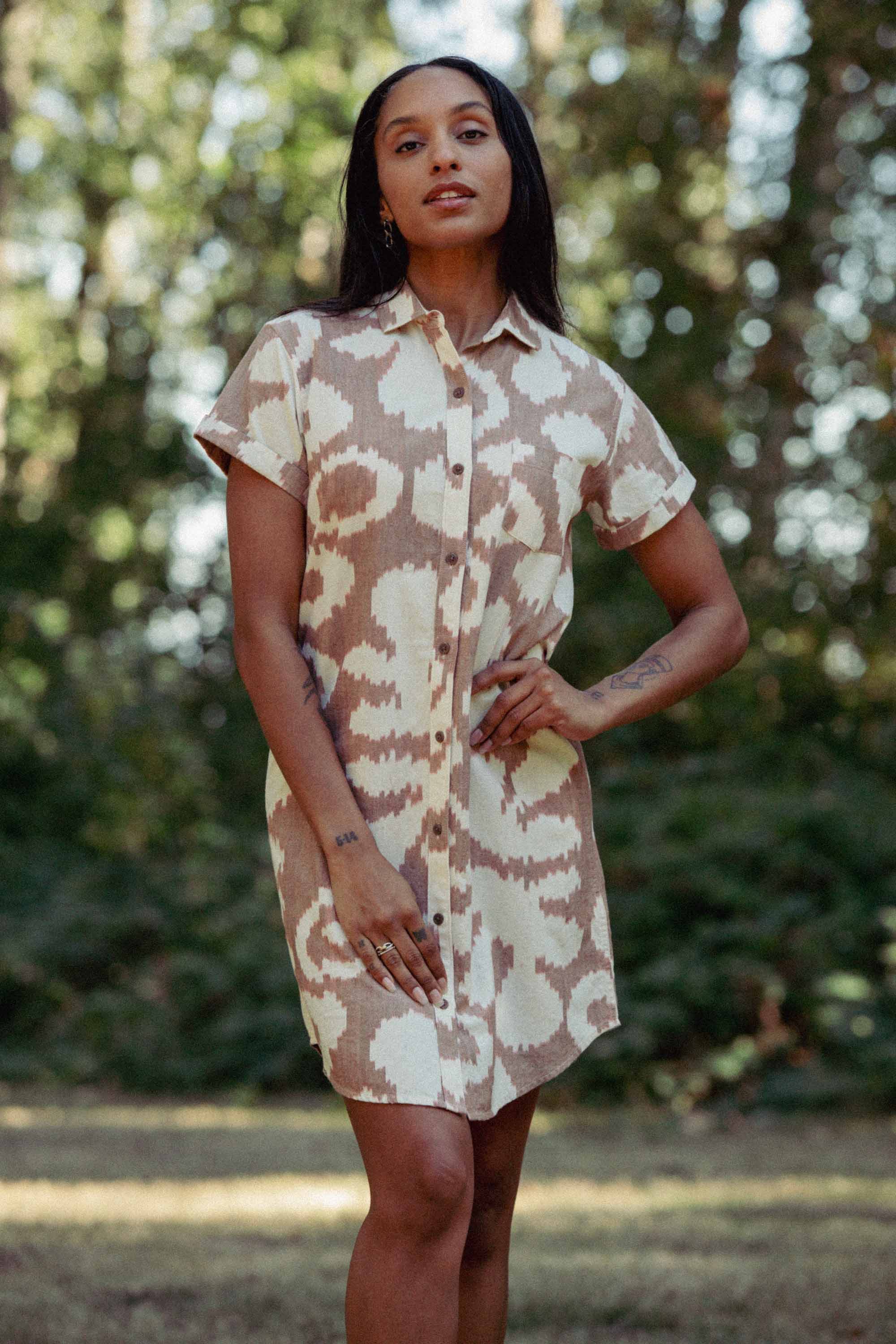A woman stands outdoors wearing the Loren Shirt Dress / Pop Leaf by Bridge & Burn—a 100% cotton, short-sleeve button-front shirt dress with a beige and white abstract pattern. She poses with one hand on her hip among trees and greenery.