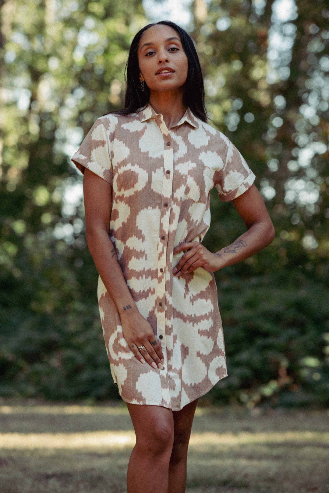 A woman stands outdoors wearing the Loren Shirt Dress / Pop Leaf by Bridge & Burn—a 100% cotton, short-sleeve button-front shirt dress with a beige and white abstract pattern. She poses with one hand on her hip among trees and greenery.