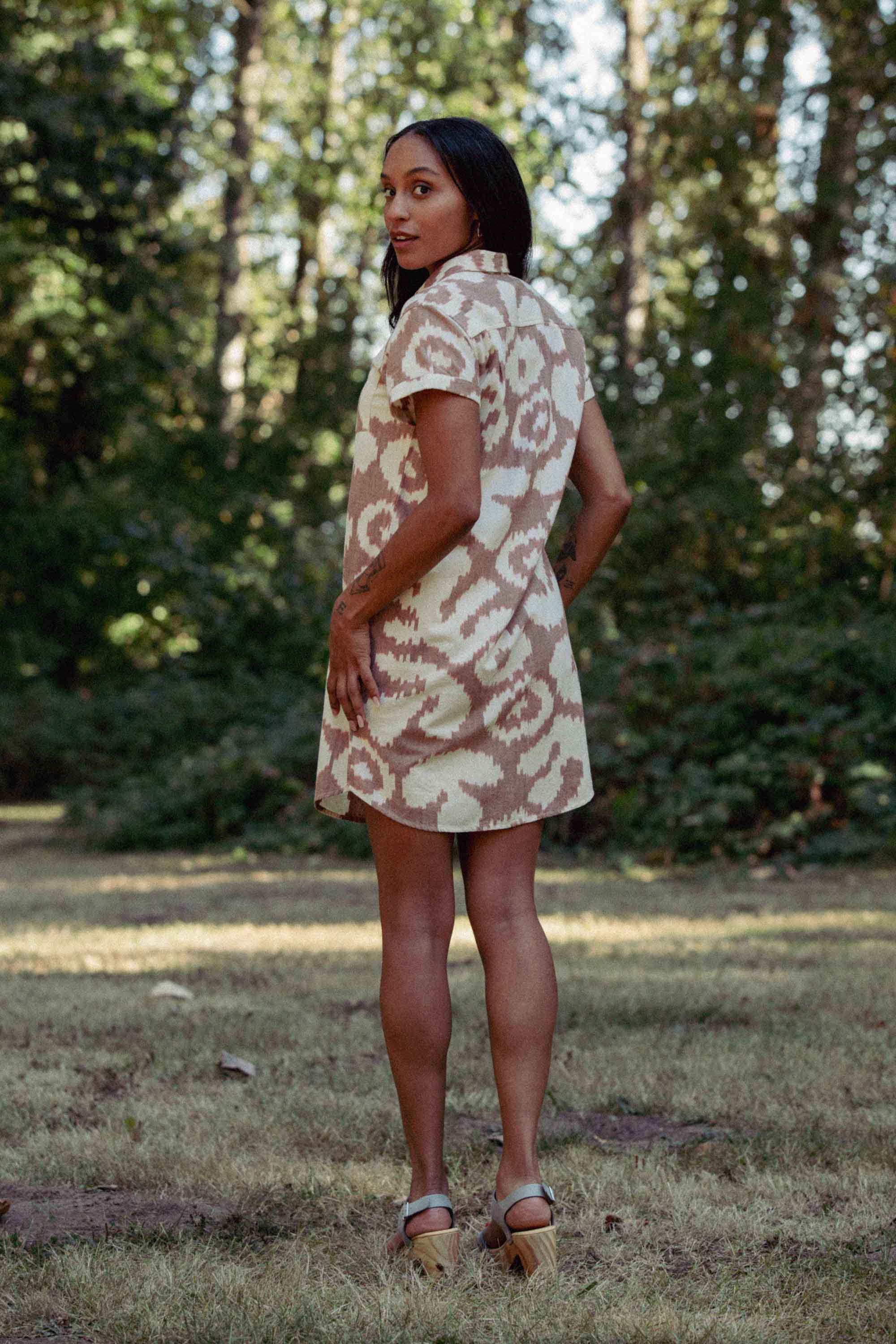 A woman stands on grass, looking over her shoulder, wearing the Bridge & Burn Loren Shirt Dress in Pop Leaf with light heeled sandals. Trees and greenery form the backdrop.