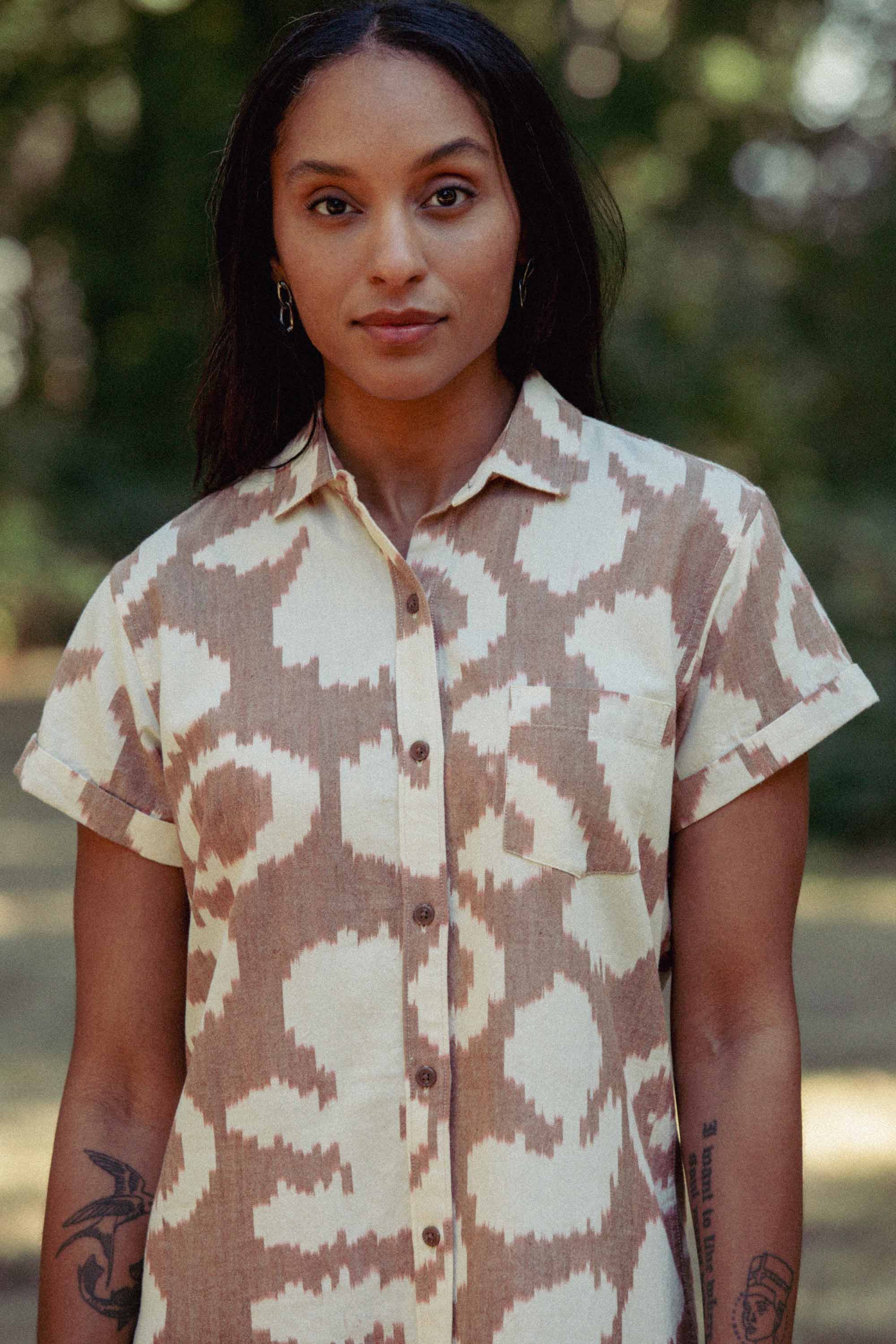 A person with long dark hair stands outdoors in the Bridge & Burn Loren Shirt Dress / Pop Leaf, a beige and white abstract print button front dress made from 100% cotton. Visible tattoos decorate their arms against a softly blurred green background.