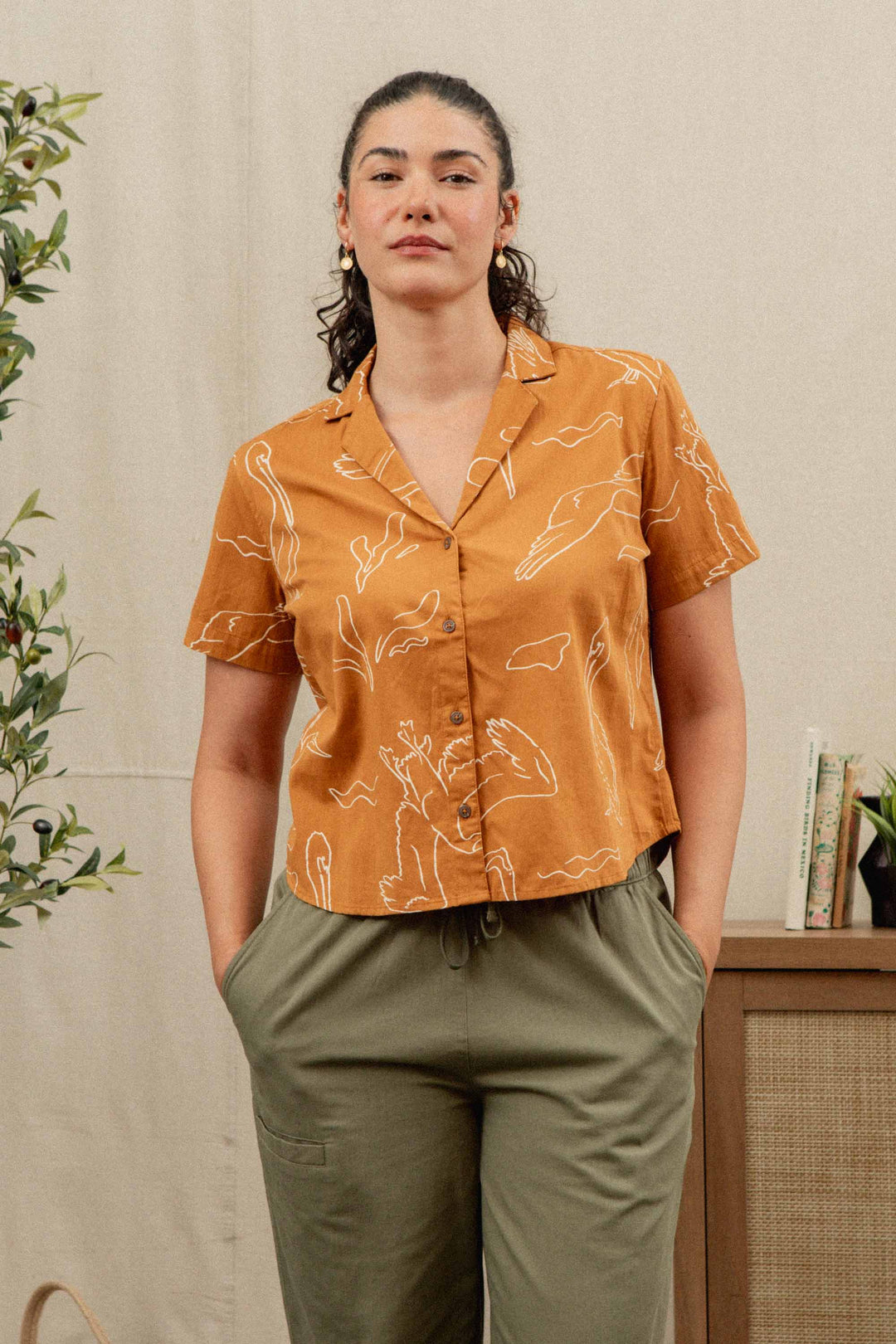 A woman with dark curly hair in a ponytail wears the Bridge & Burn Mila Cropped Shirt in Shoreflight, an orange relaxed fit short-sleeve button-up with a white abstract line pattern, paired with olive green pants indoors, hands in her pockets.