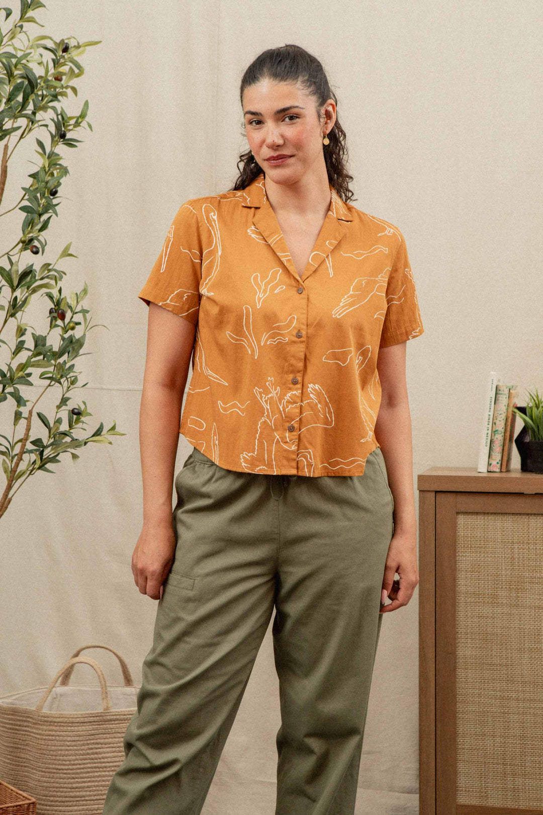 A woman with dark hair pulled back wears the Bridge & Burn Mila Cropped Shirt in Shoreflight—an orange, relaxed-fit, short-sleeved top with white line patterns—paired with olive green pants indoors by a plant and cabinet with books.