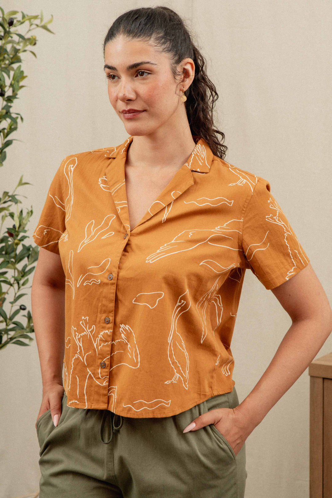 A woman with dark hair tied back wears the Bridge & Burn Mila Cropped Shirt in Shoreflight—an organic cotton, rust-colored short-sleeve button-up with white animal line drawings—and green pants, standing with hands in her pockets against a neutral background.