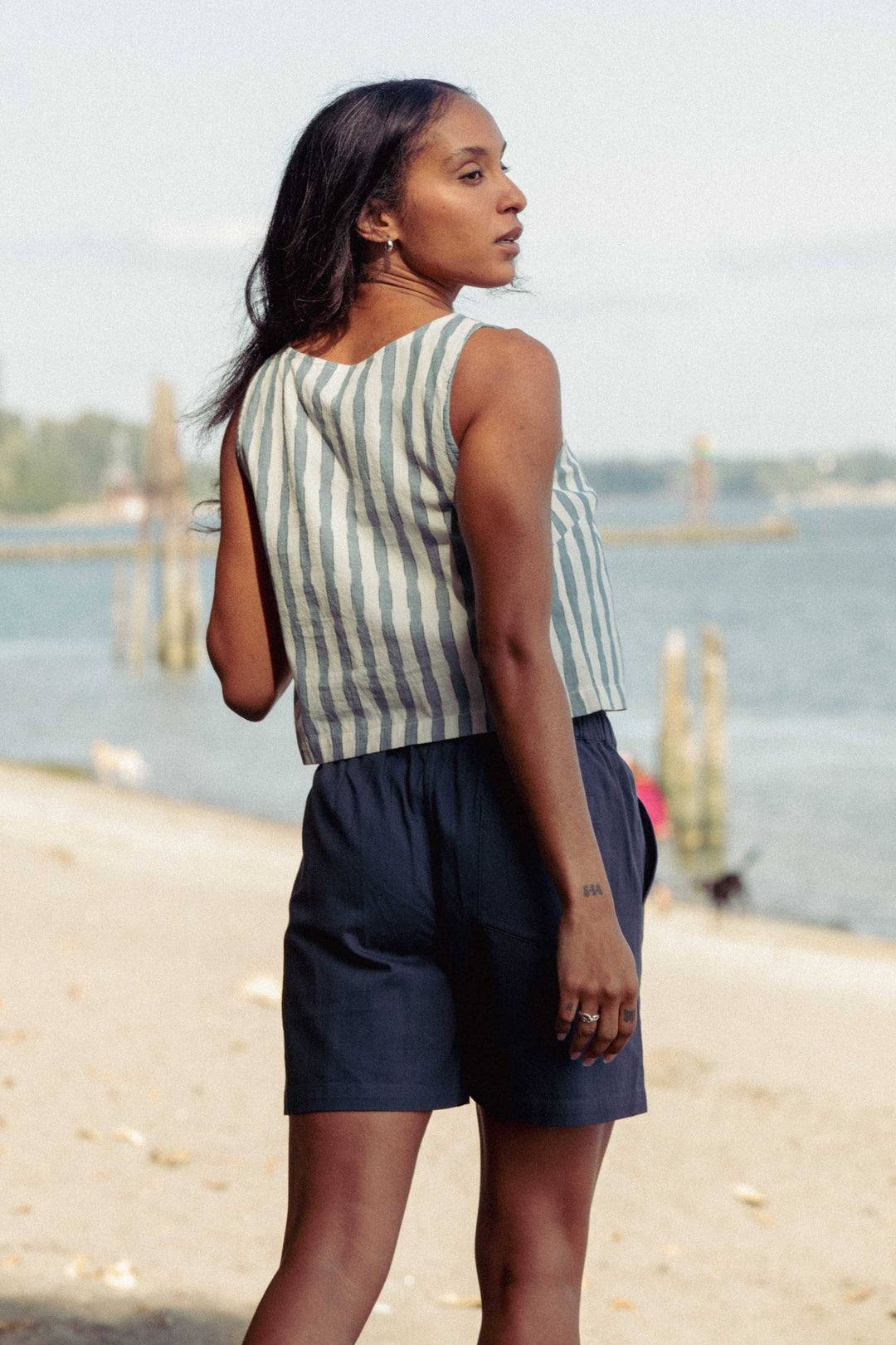 A woman stands on a sandy beach facing away, looking over her shoulder. She wears the Bridge & Burn Sadie Tank in Beach Stripe and dark shorts. Water, wooden posts, and distant trees make up the background.
