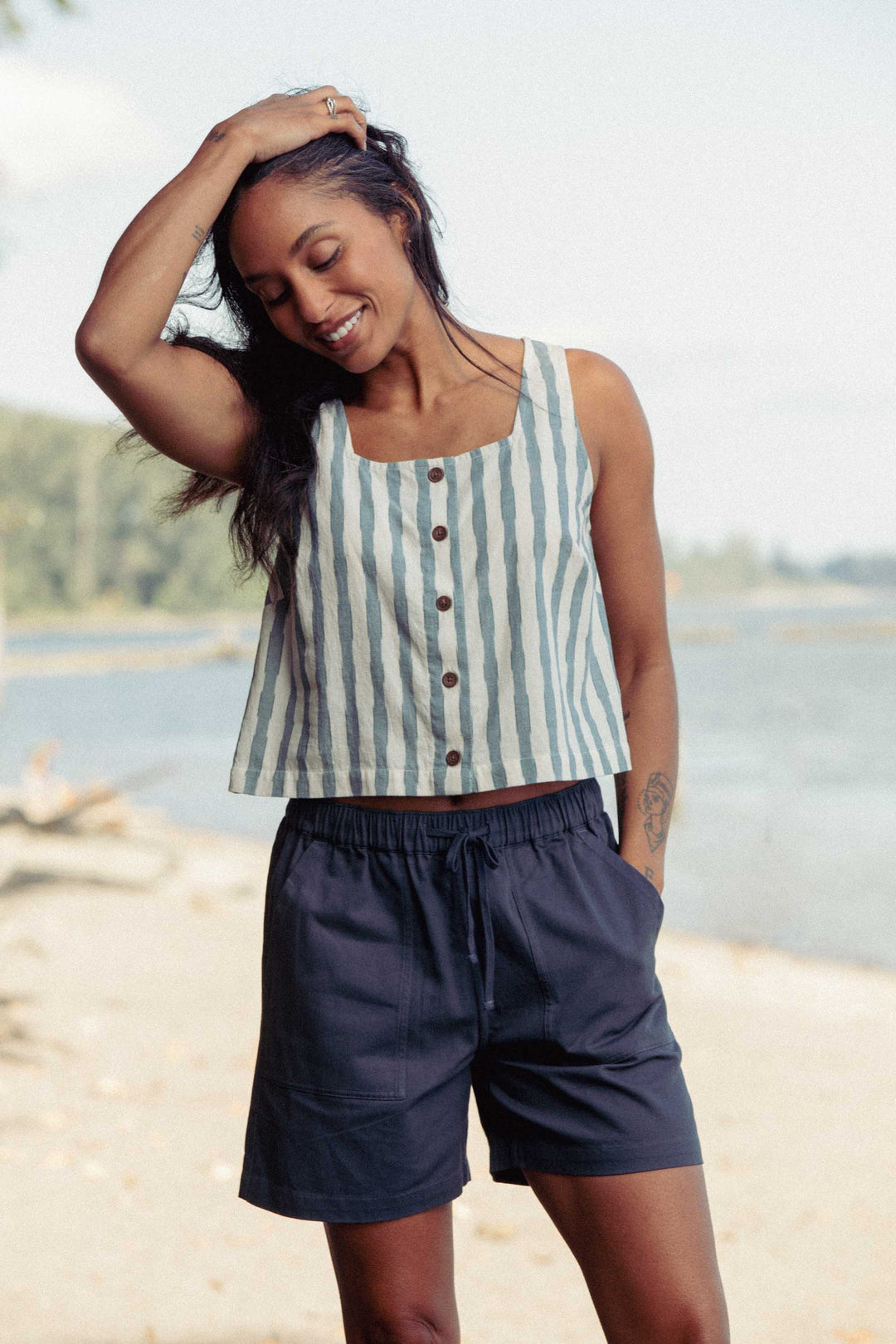 A smiling woman stands outdoors by the water wearing the Bridge & Burn Sadie Tank in Beach Stripe and dark shorts, with one hand in her pocket and the other touching her head. Trees and a blurred shoreline are in the background.